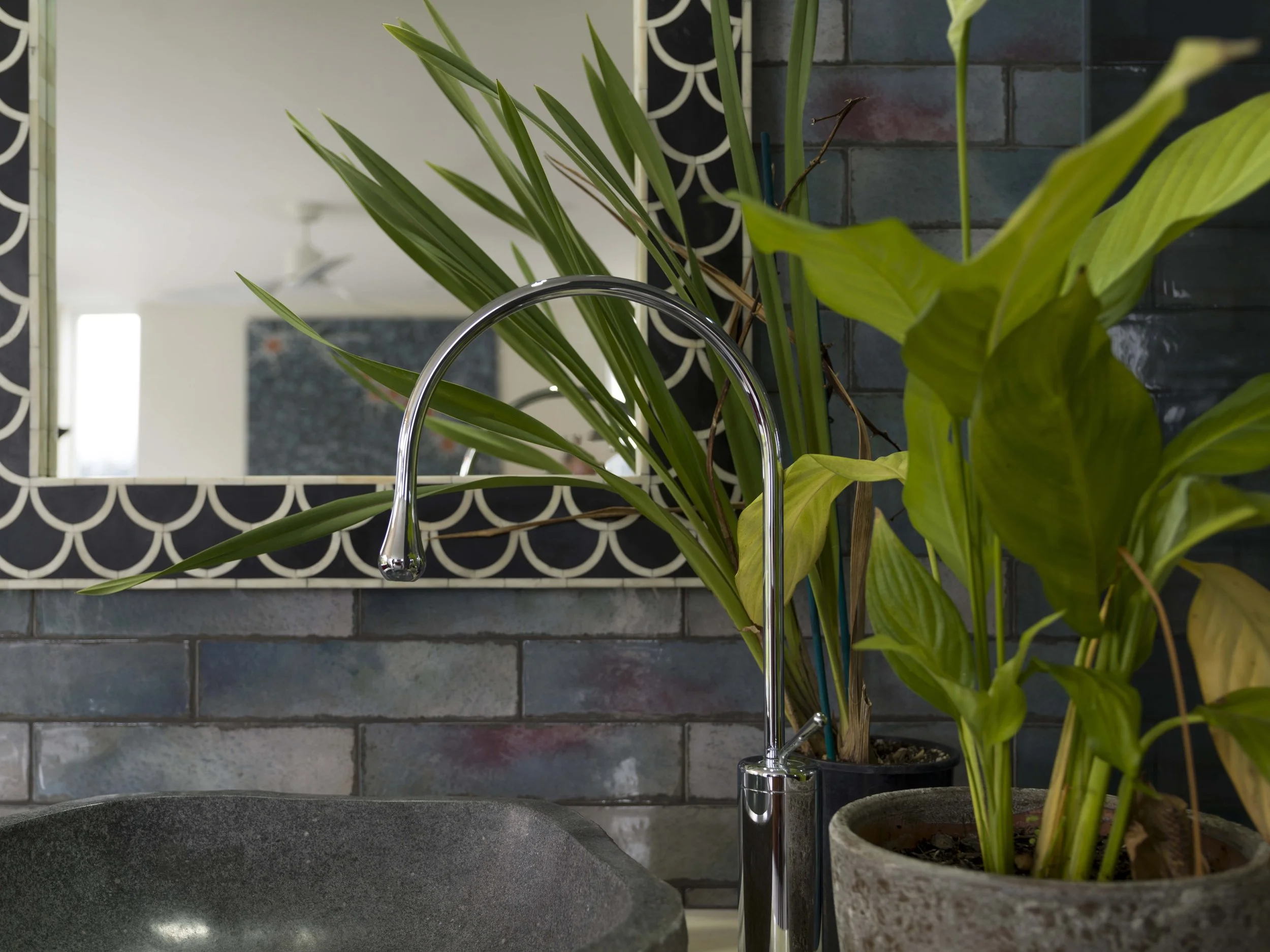 Close-up of a kitchen sink with a curved silver faucet, surrounded by green plants and a rectangular mirror with a black and white scalloped pattern frame, against a dark brick wall.