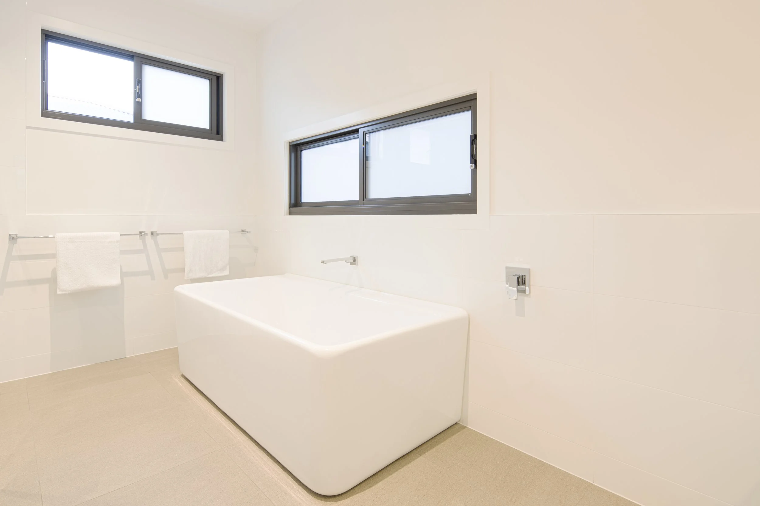 A modern bathroom with a white bathtub, two windows with black frames, white towels hanging on a rail, and a beige tiled floor.