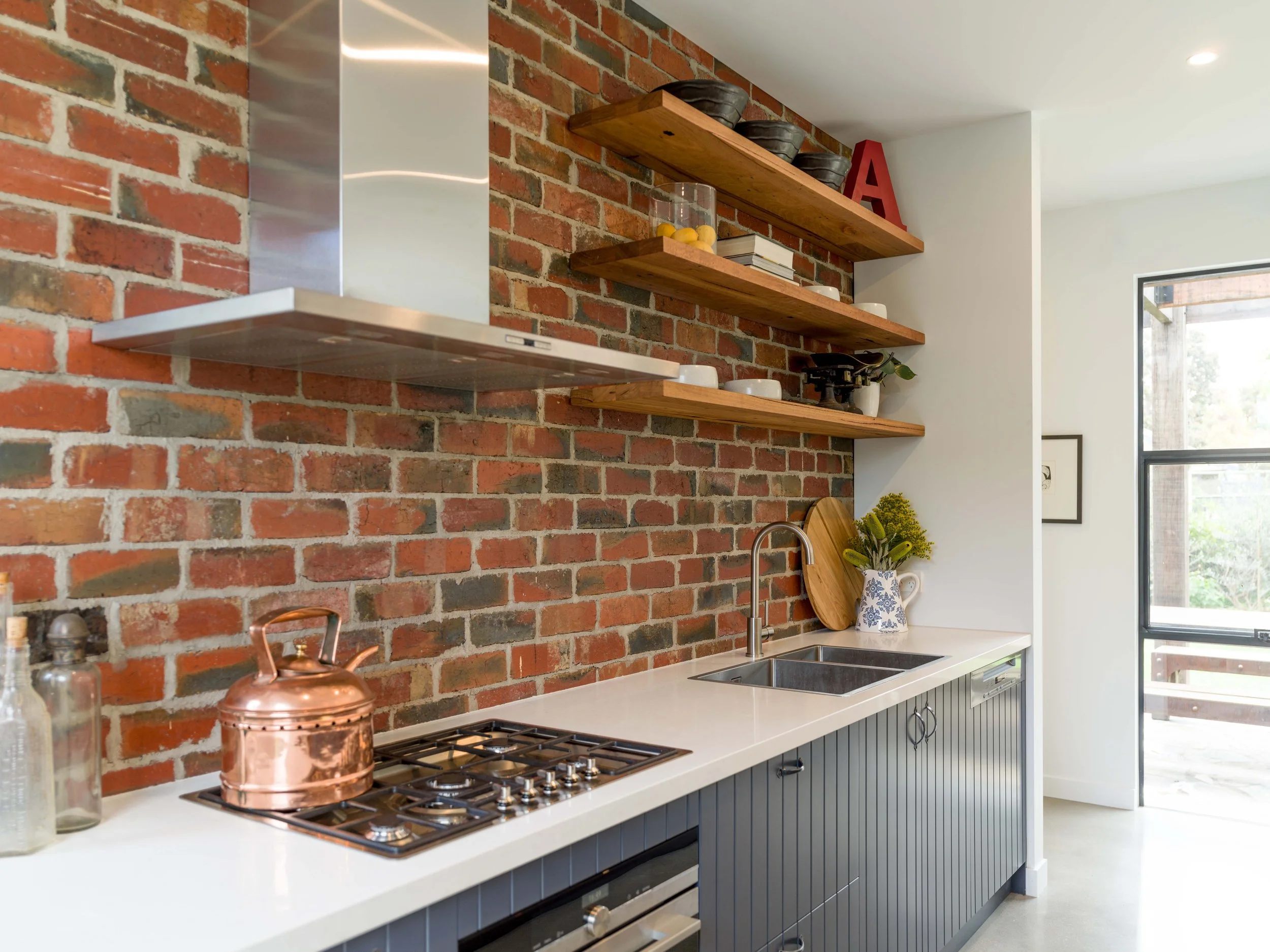 Modern kitchen with brick wall, wooden shelves holding bowls and books, white countertop, stainless steel range hood, black cabinets, and a window with a view of trees outside.