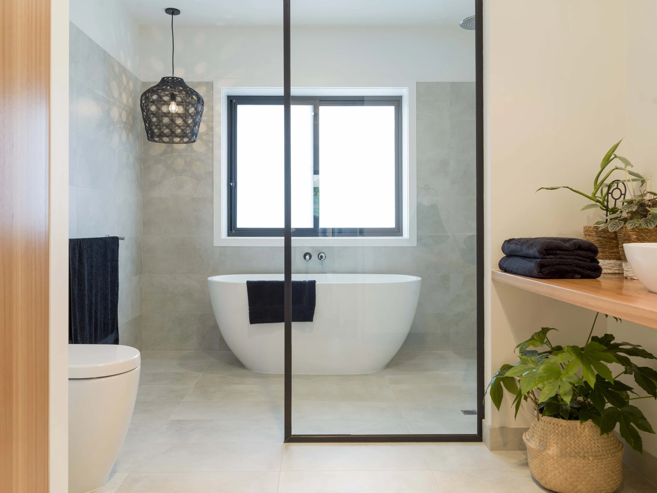 Modern bathroom with a free-standing bathtub below a large window, black towel on a rack, wicker basket with green plants on a wooden shelf, black pendant light, glass shower enclosure, and beige tiled walls and floor.