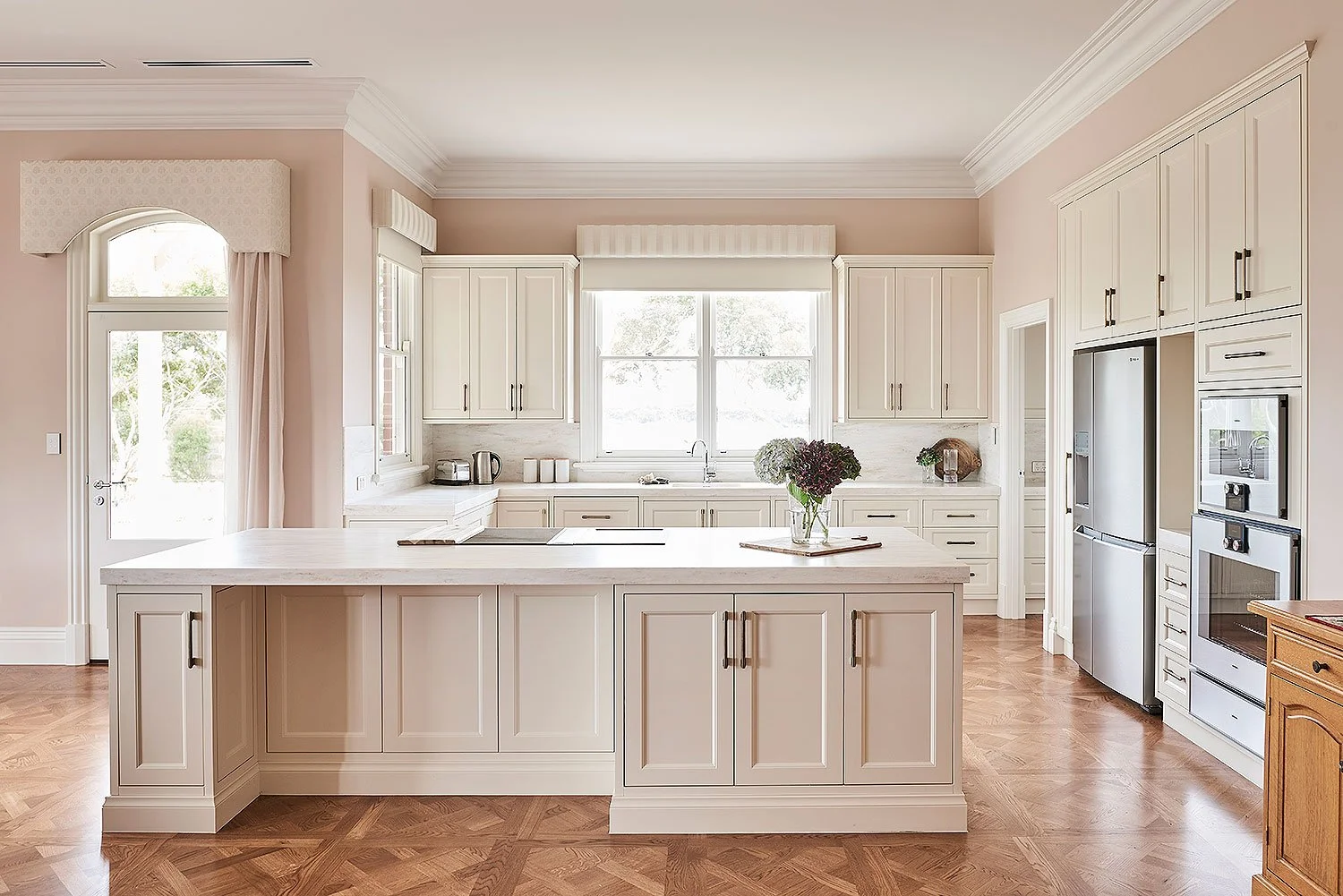 Bright kitchen with white cabinets, a central island, stainless steel refrigerator and oven, large window over the sink, wooden floor, and soft pink walls.