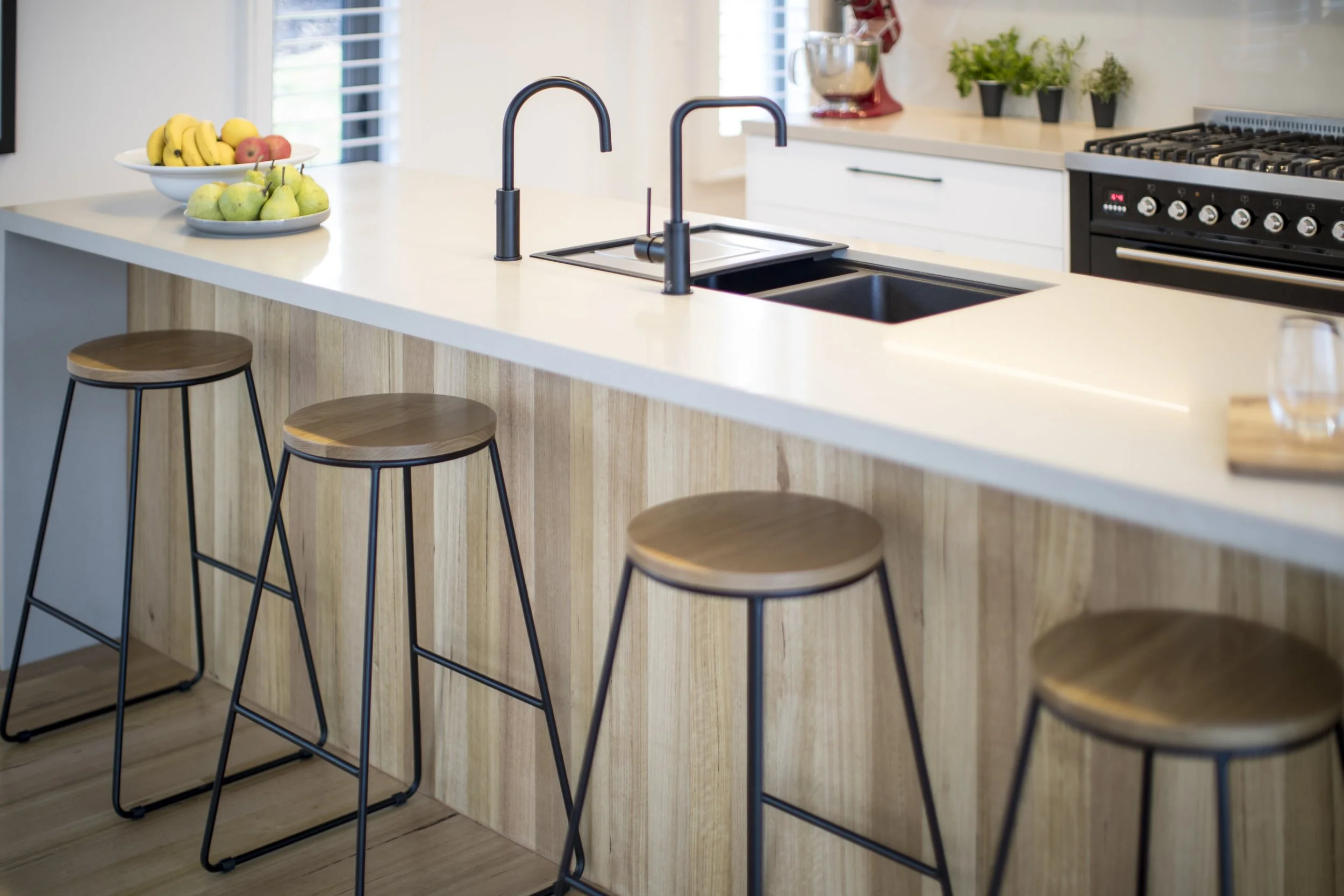 Kitchen island with a white countertop, three wooden bar stools with black metal legs, a black faucet, and a double sink. In the background, there are bowls of bananas and pears on the countertop, a window with blinds, and a few potted plants near th