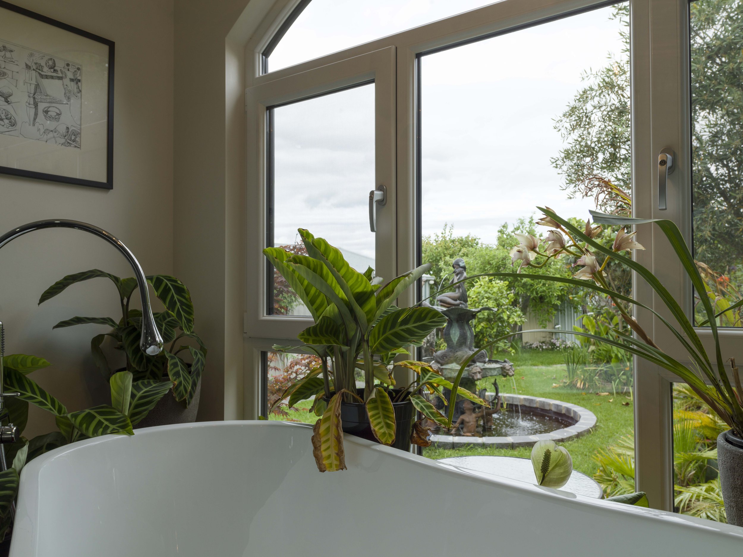 Indoor view of a bathtub with potted plants near a large window revealing a backyard garden with trees, a frog statue, and a small pond, with a framed drawing on the wall.