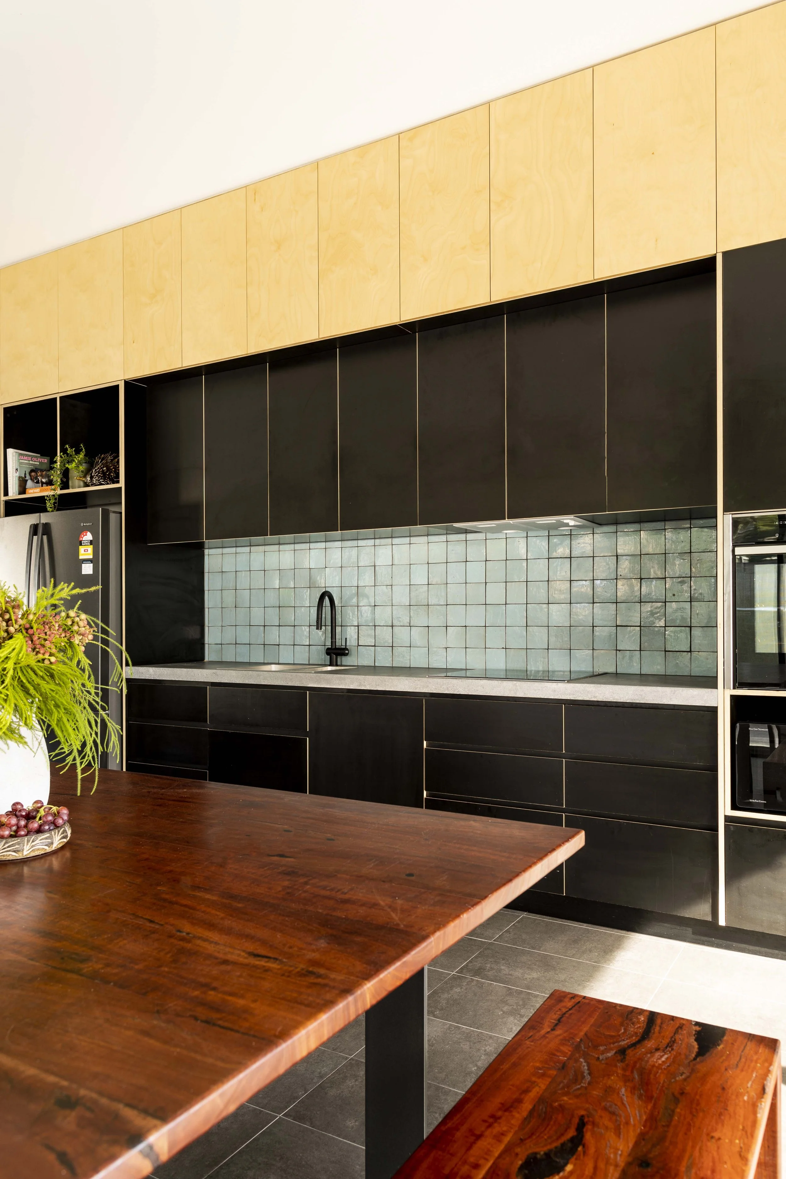 Modern kitchen with black and wood cabinets, light blue tile backsplash, and a wooden table with a potted plant and grapes.