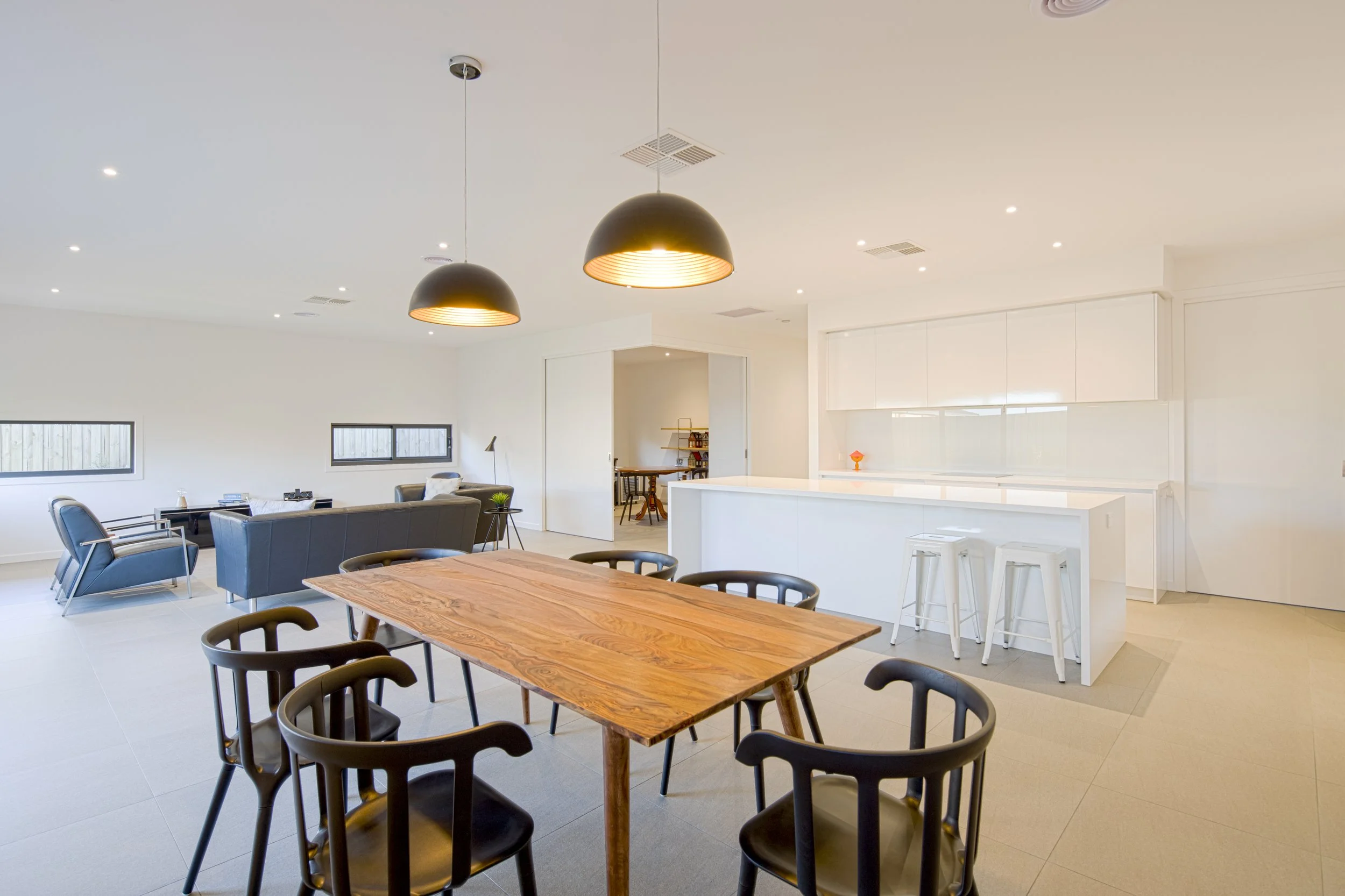 Modern open-concept living and dining area with a wooden dining table, black chairs, a sitting area with gray sofas, and black pendant lights hanging from the ceiling.