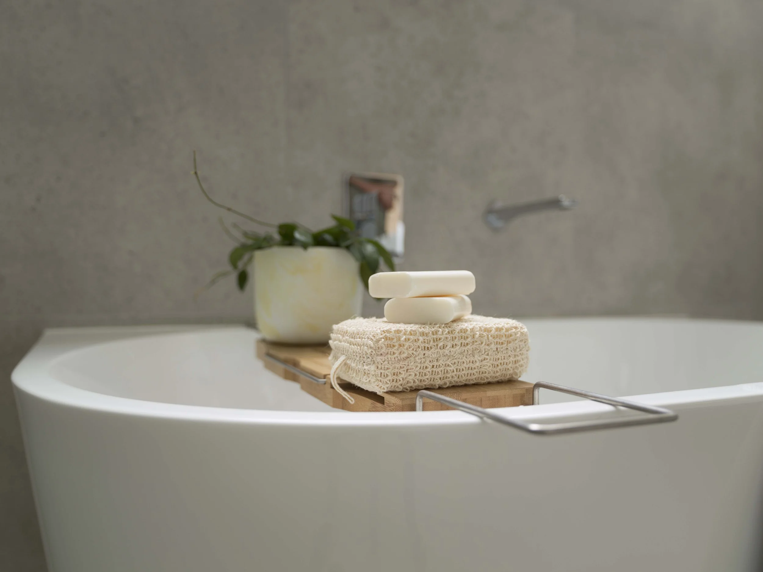 A stack of three white soap bars placed on a knitted beige cloth on a wooden tray, with a potted plant and a framed photo in the background, against a gray wall.