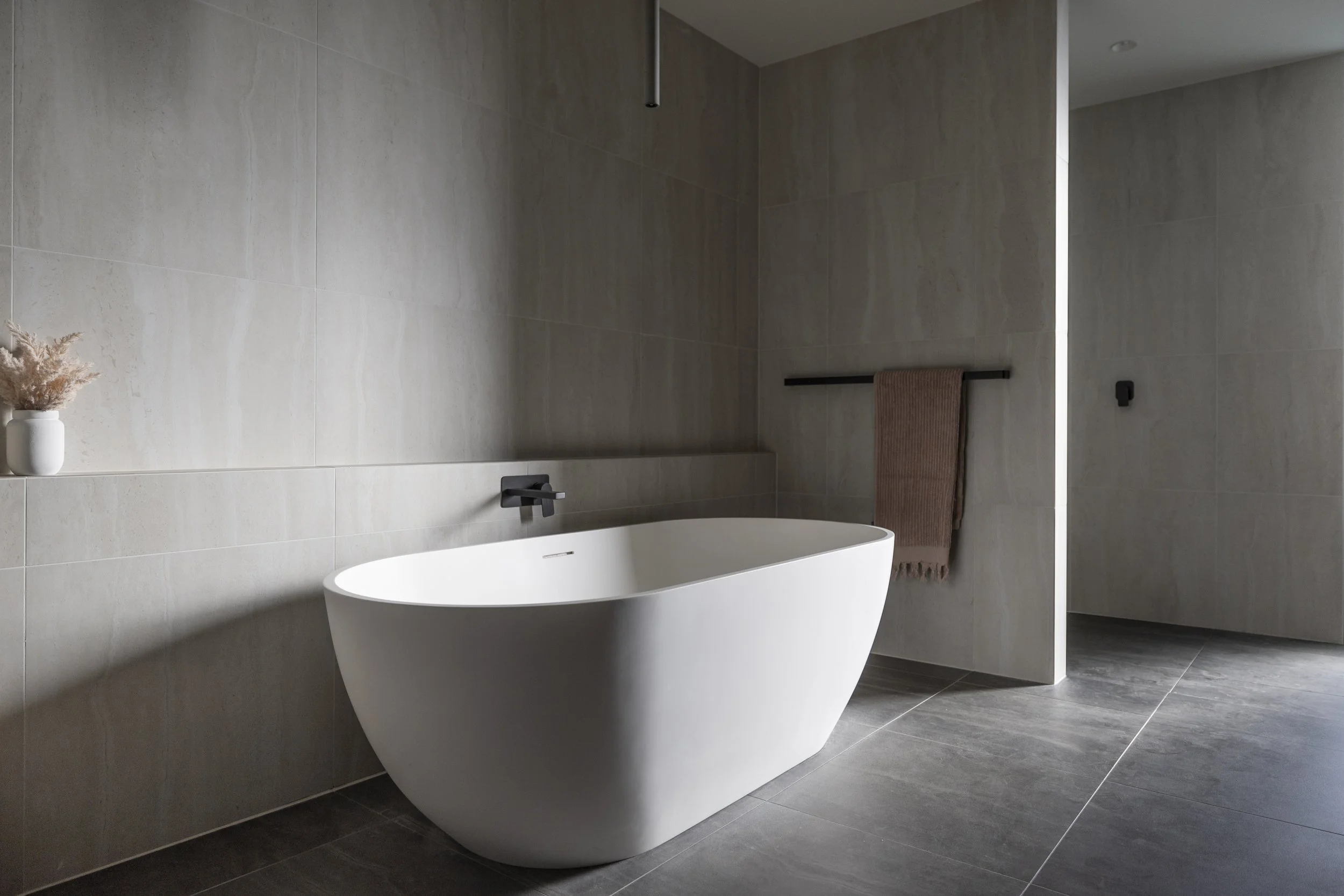 Modern bathroom with a white freestanding bathtub, gray tiled walls, a black towel rack with a brown towel, a small decorative plant in a white vase, and a slim wall-mounted cabinet.
