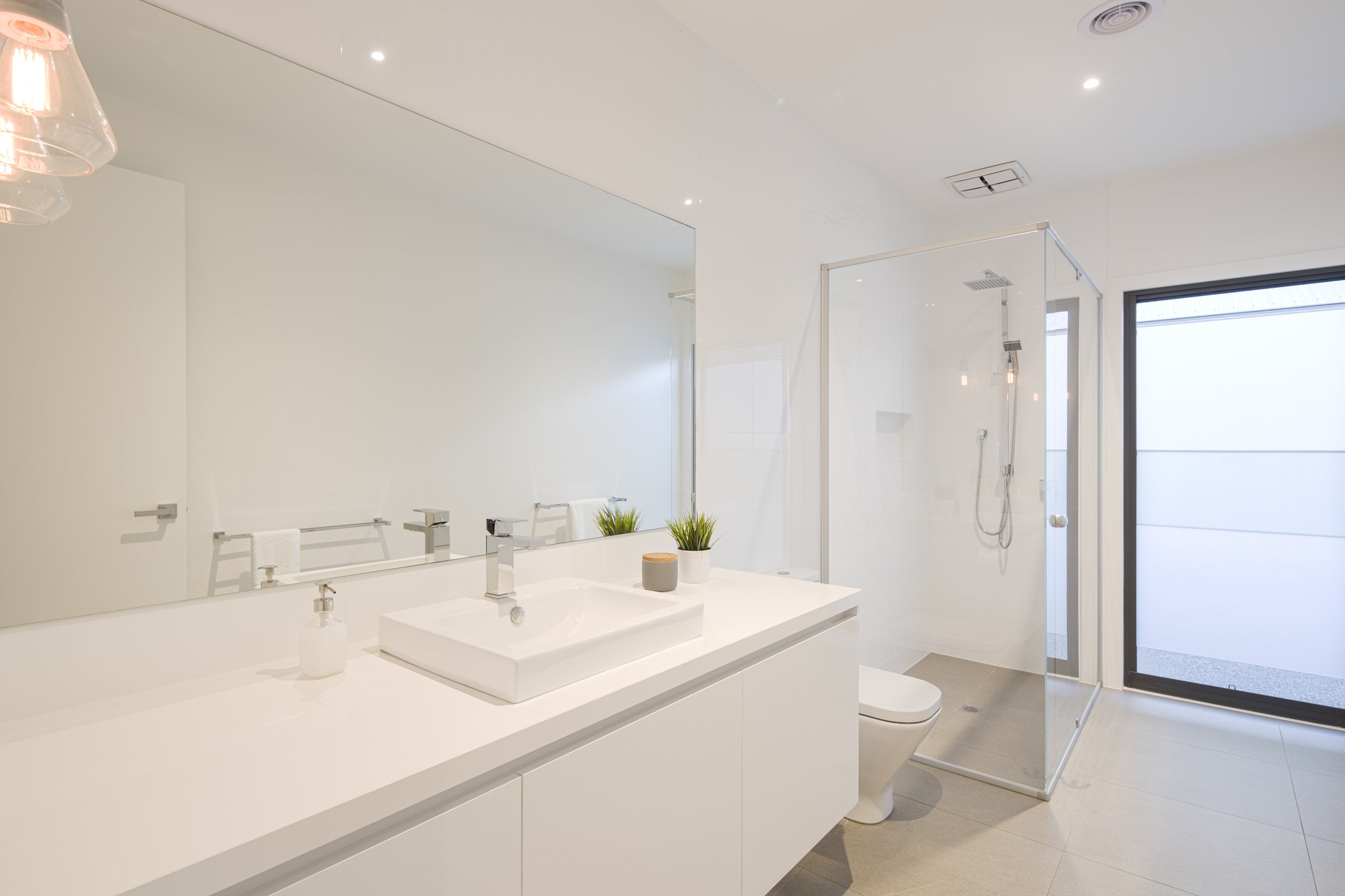 Modern white bathroom with vanity, square sink, large mirror, plants, glass-enclosed shower, and toilet, with natural light from a large window.