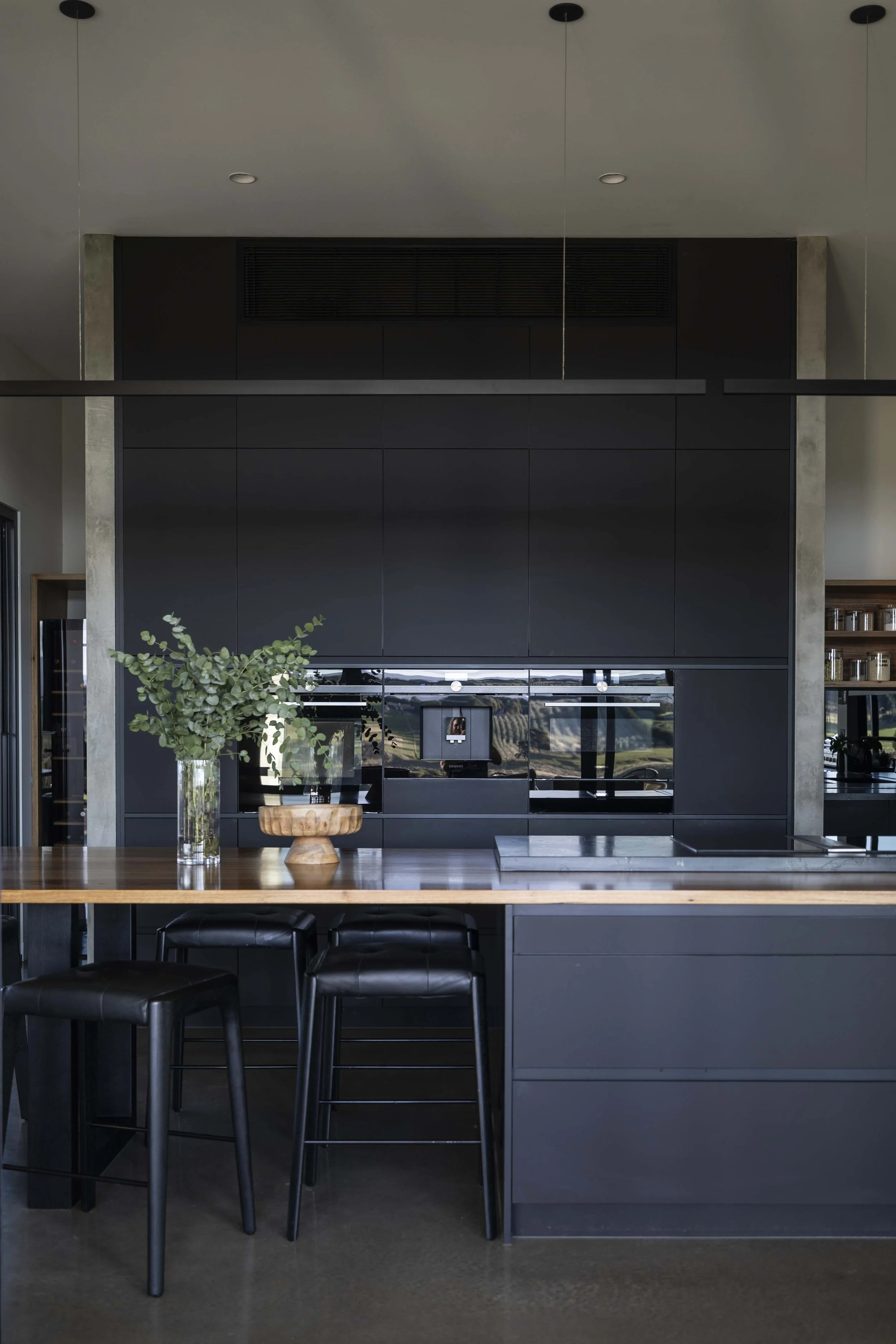 Modern kitchen with black cabinetry, kitchen island, and wooden table with black chairs. Green plant and wooden bowl on the table. Large mirrors or windows reflecting landscape view.