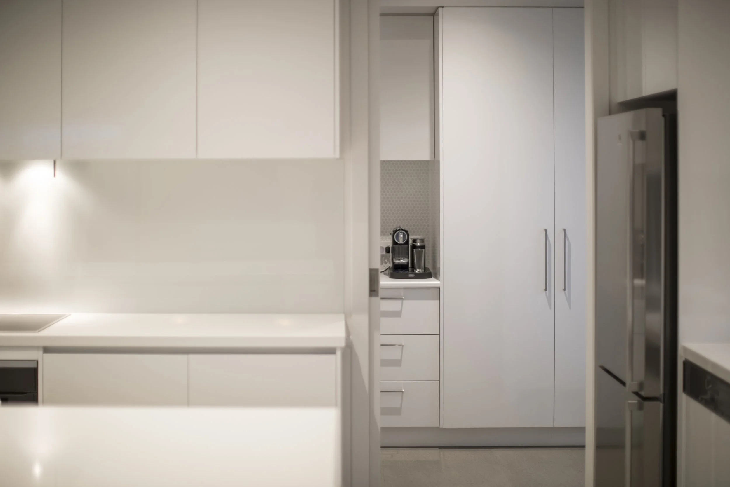 A modern white kitchen with minimalistic cabinets and a countertop. There is a coffee machine with a glass on a small shelf. The refrigerator is visible on the right side.
