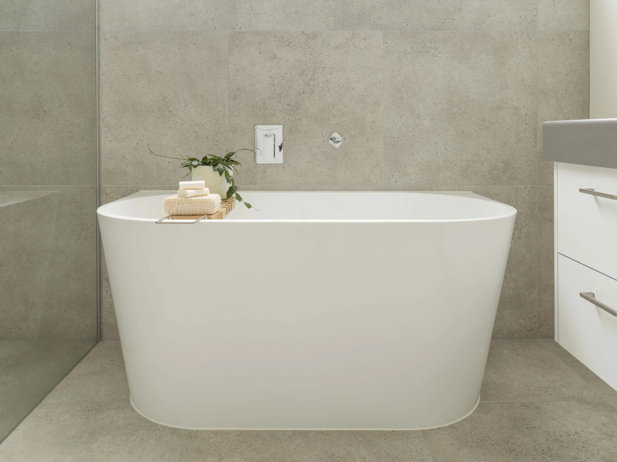 A modern bathroom with a white freestanding bathtub, a potted plant, soap, and towels on a wooden tray. The background has beige tiled walls and a corner with a glass shower partition.