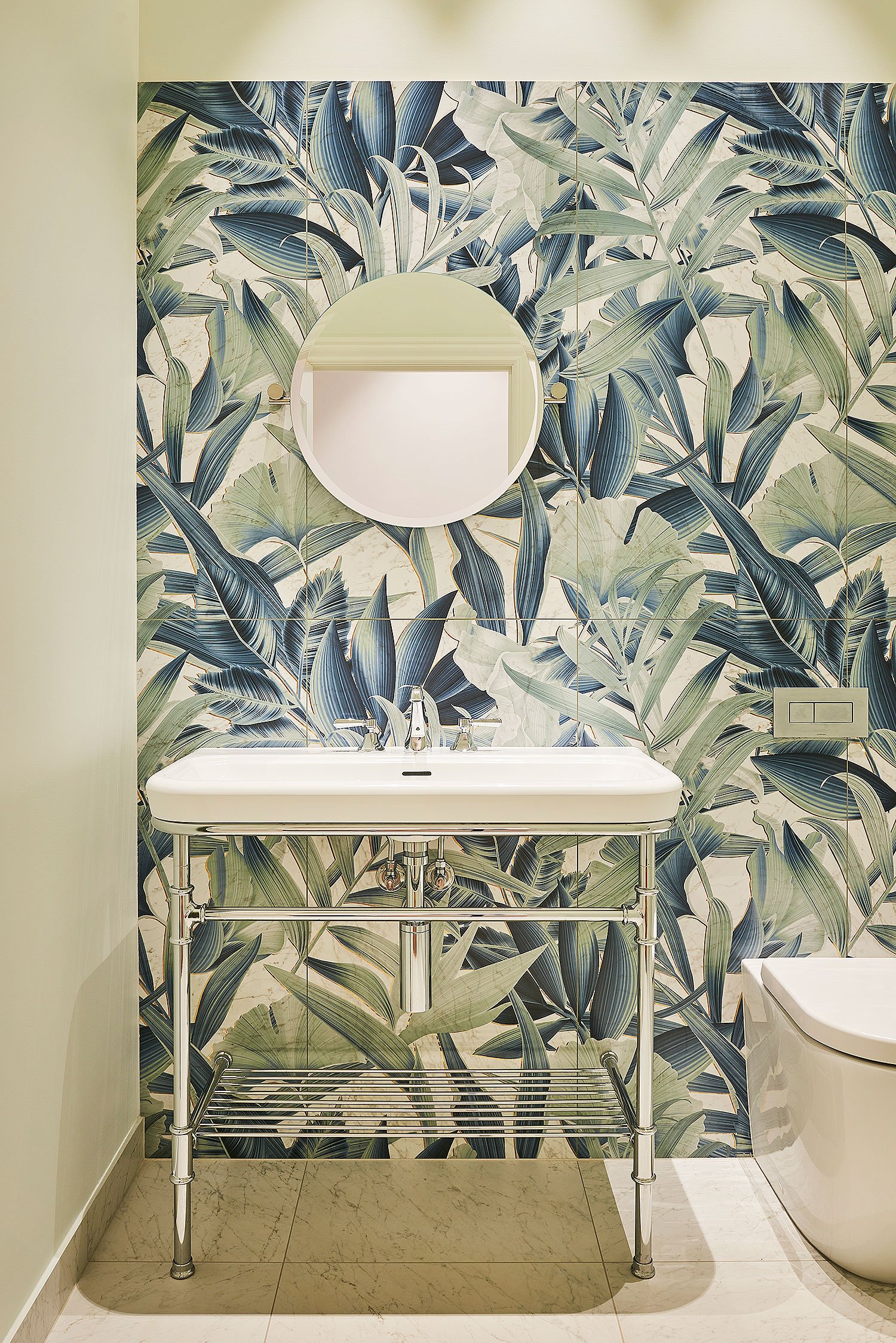 Bathroom with a floral wallpaper featuring large blue and green leaves, a white oval mirror, a vintage-style white sink with chrome fixtures, and a modern white toilet on a beige tiled floor.