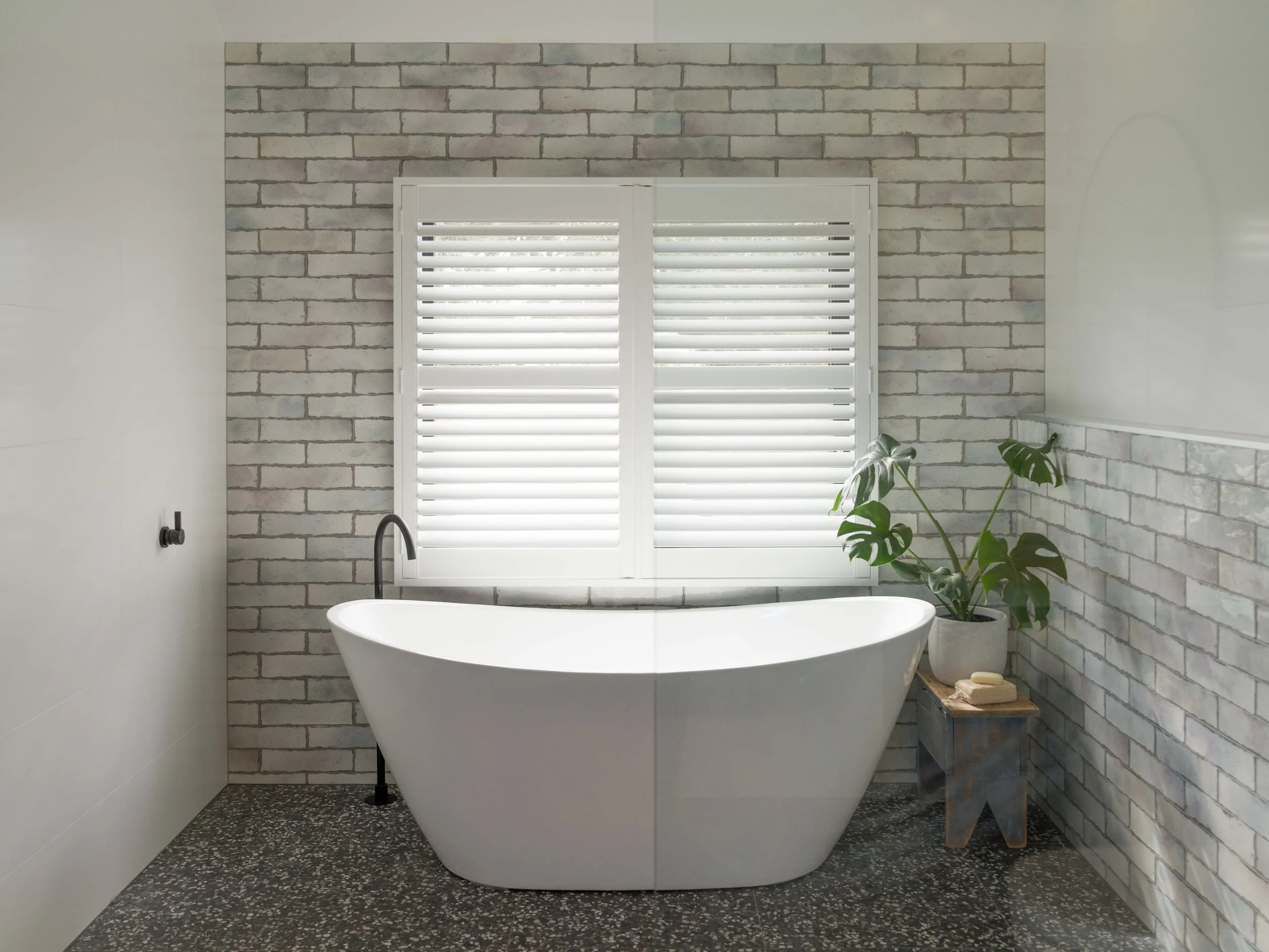 A modern bathroom with a white freestanding bathtub in front of a brick wall and a window with white shutters. To the right, a small wooden stool holds towels and bath accessories. A green potted plant is positioned beside the stool.