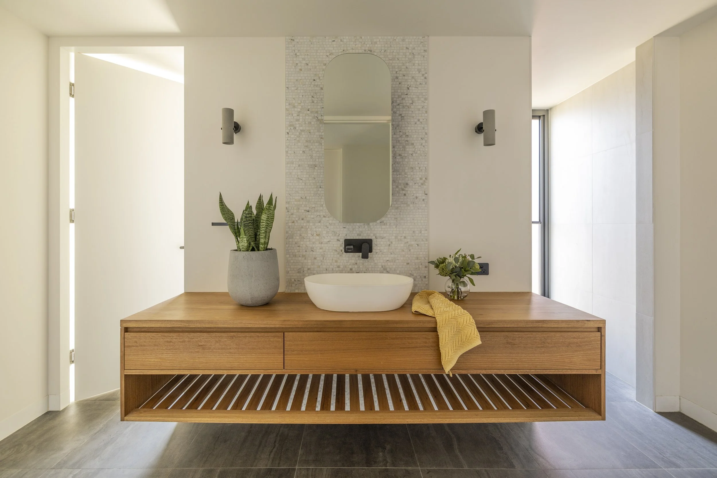 Modern bathroom with a wooden vanity, oval mirror, plants, and decorative wall lights.
