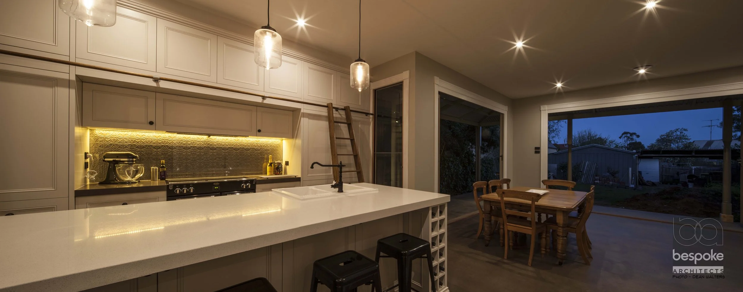 Modern kitchen with white island, hanging pendant lights, dining table with chairs, sliding glass door, and outdoor view at dusk.