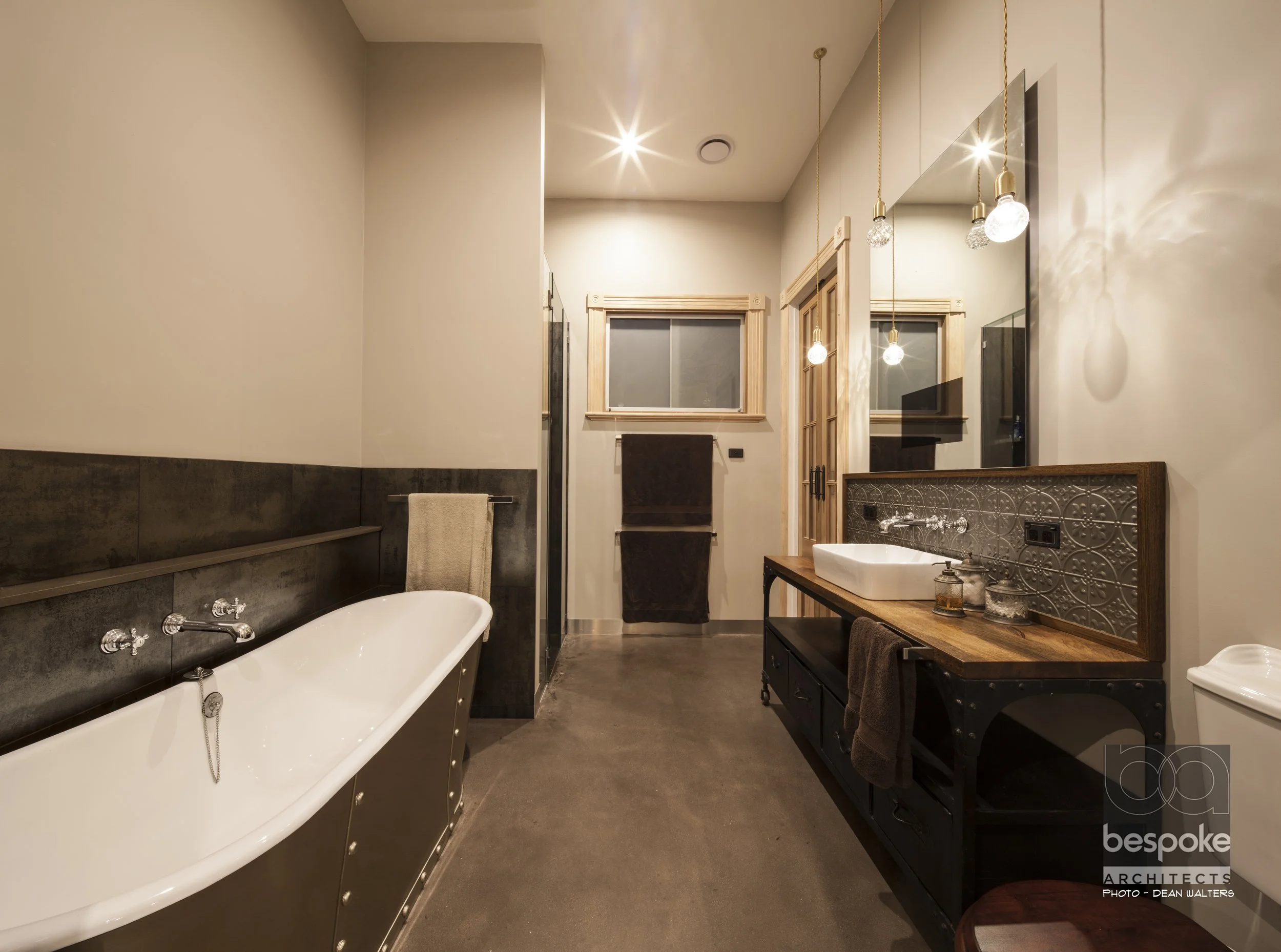 Modern bathroom with a freestanding bathtub on the left, dark tiled walls, a wooden vanity with a white sink on the right, and three pendant lights hanging from the ceiling. There is a mirror above the vanity and a window with wooden trim on the back