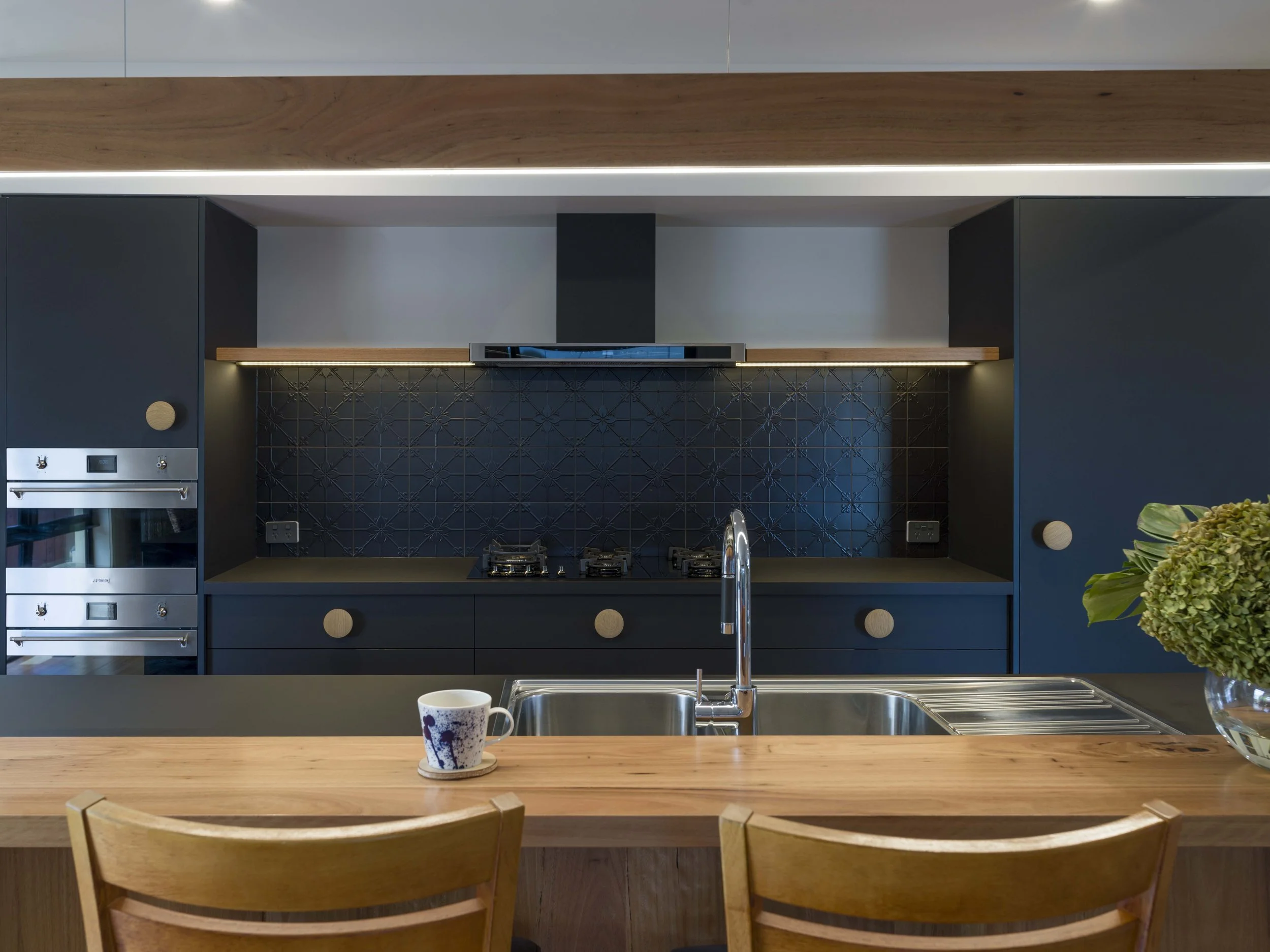 Modern kitchen with black cabinets, a gas stove, a black and white patterned tile backsplash, and a wooden kitchen island with chairs. A blue and white mug is on the island, and a potted plant is on the right.