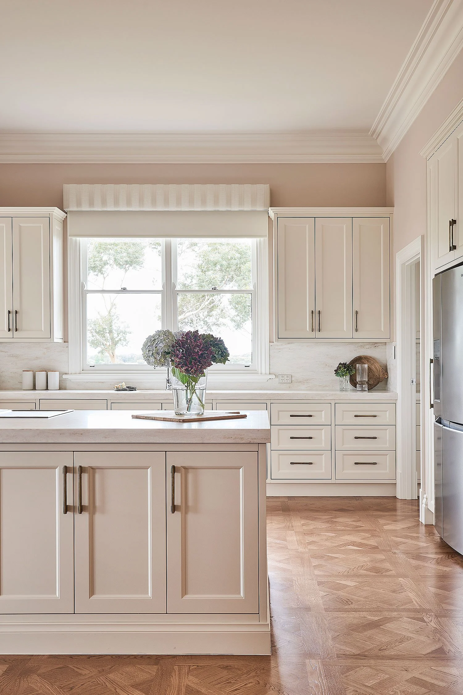 A bright kitchen with cream-colored cabinets, a large window with a window valance, a kitchen island with a flower vase, and hardwood flooring.