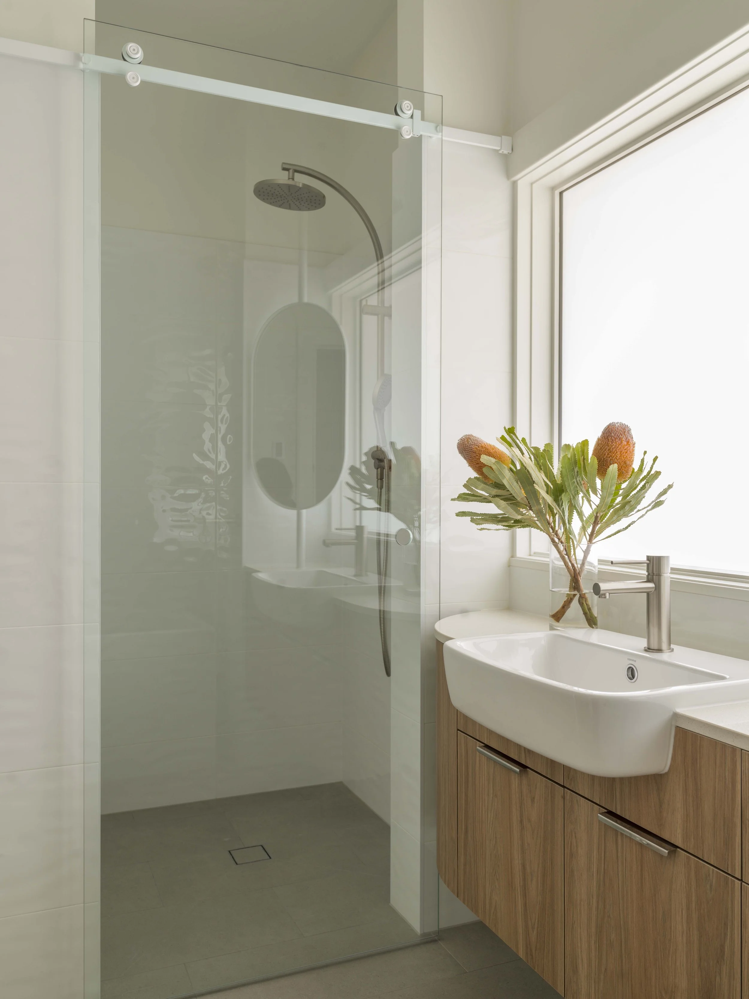 Modern bathroom with a walk-in shower with glass door, a round white sink, wooden cabinetry, a large window with a planter and green plants, and natural light filling the space.