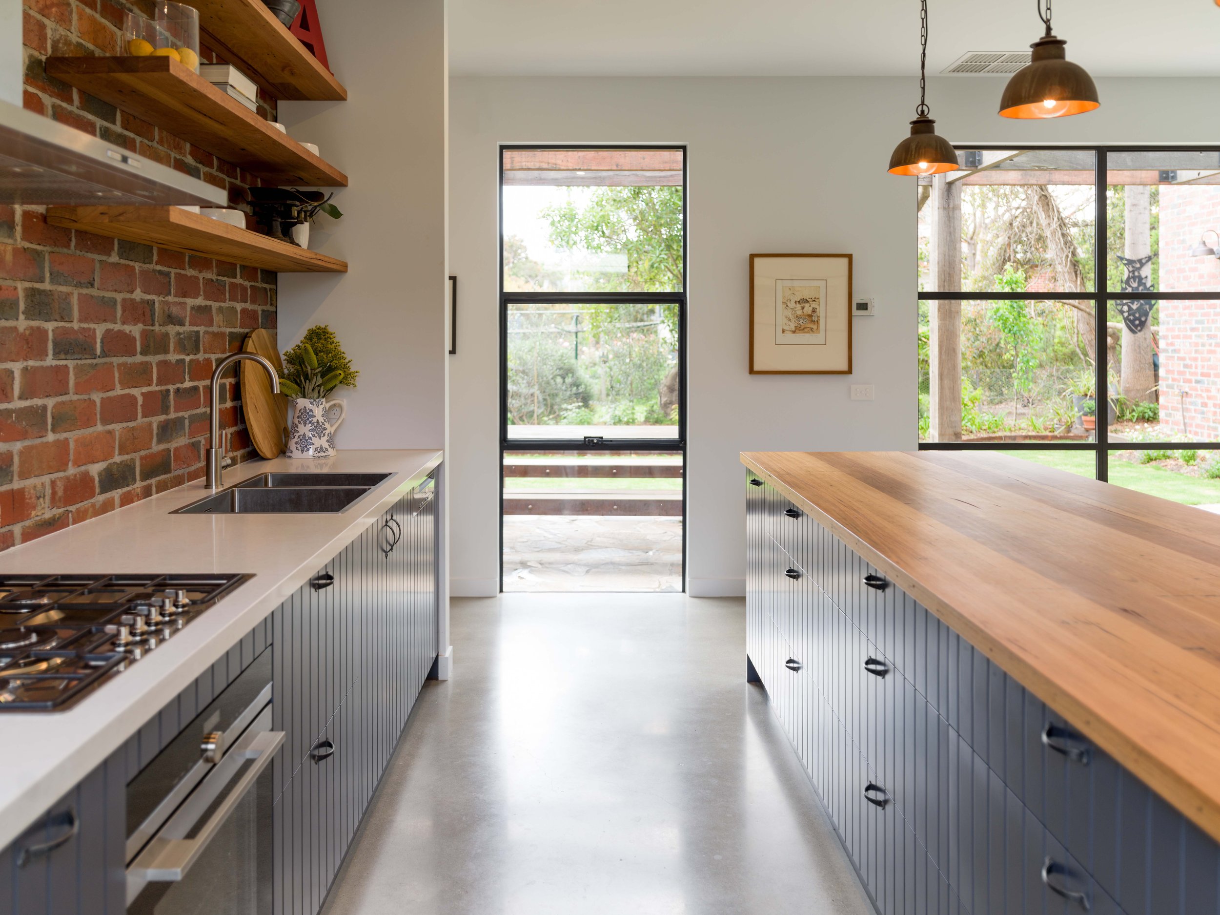 Modern kitchen with brick wall, open wooden shelves, and large windows showing outdoor greenery.