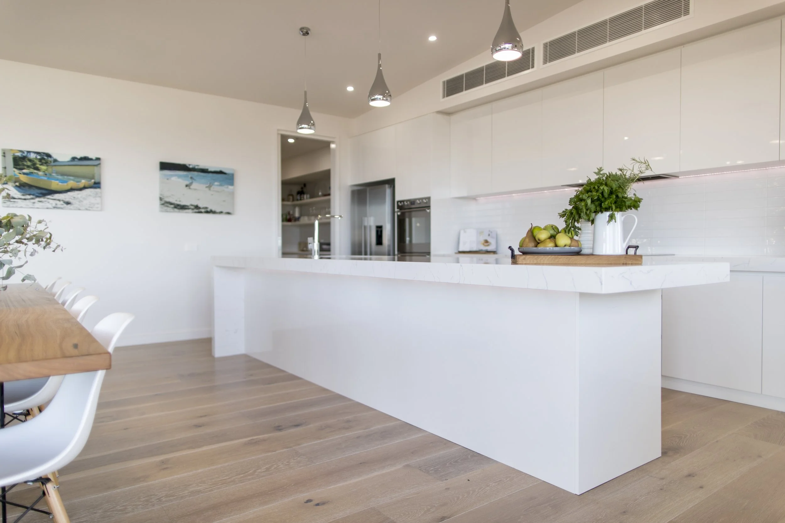 Modern kitchen with white cabinets, a white marble island, hanging pendant lights, and a wooden floor. There is a basket of pears and a potted plant on the island, and a dining table with white chairs partially visible on the left.
