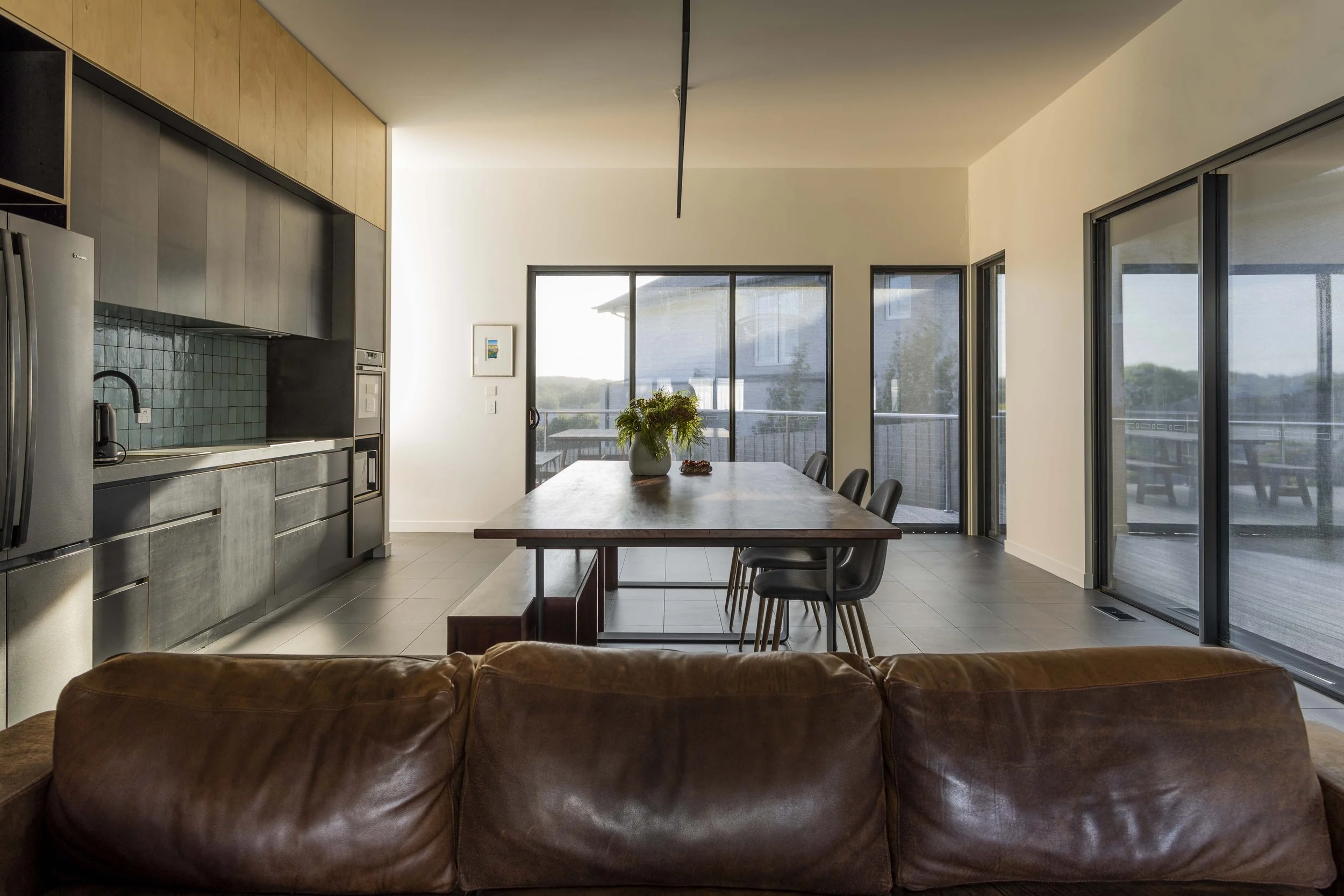 Modern kitchen and dining area with large windows, a wooden dining table, black chairs, and a sliding glass door leading to an outdoor balcony.