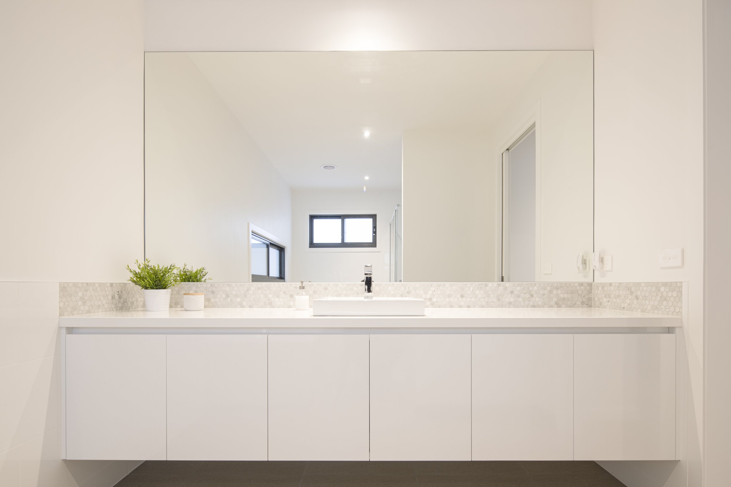 Modern bathroom with a large mirror, white vanity, potted plant, and window in the background.