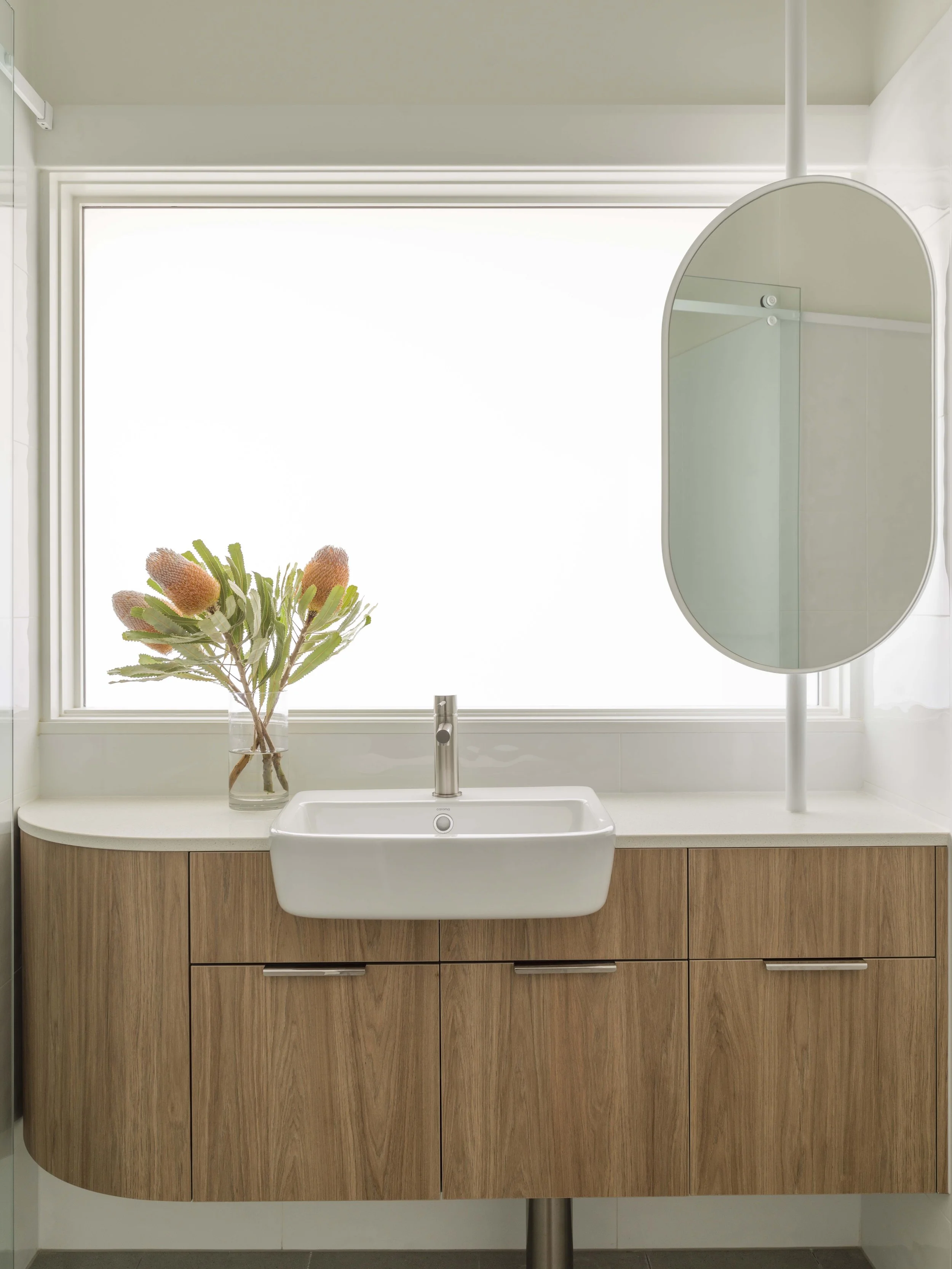 A bathroom vanity with a white sink, a wooden cabinet, a faucet, and a large window with a plant placed in front of it. There is a decorative mirror above the sink.