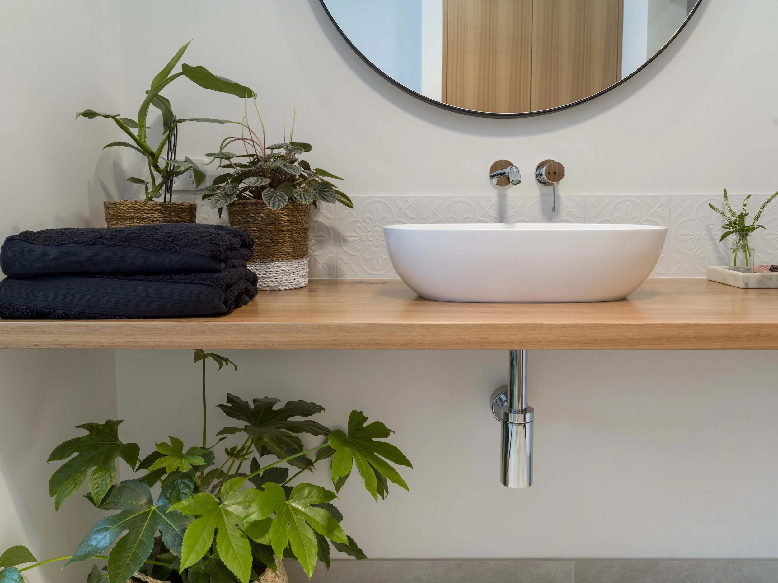 A bathroom countertop with potted plants, rolled black towels, a white vessel sink, a circular mirror, and a wooden cabinet reflected in the mirror.