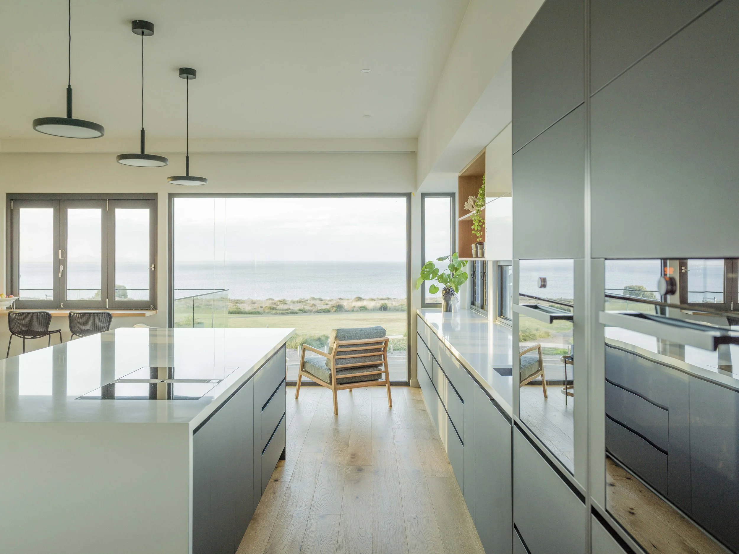 Modern kitchen with large windows overlooking a seascape, featuring white countertops, dark cabinetry, and minimalist decor.