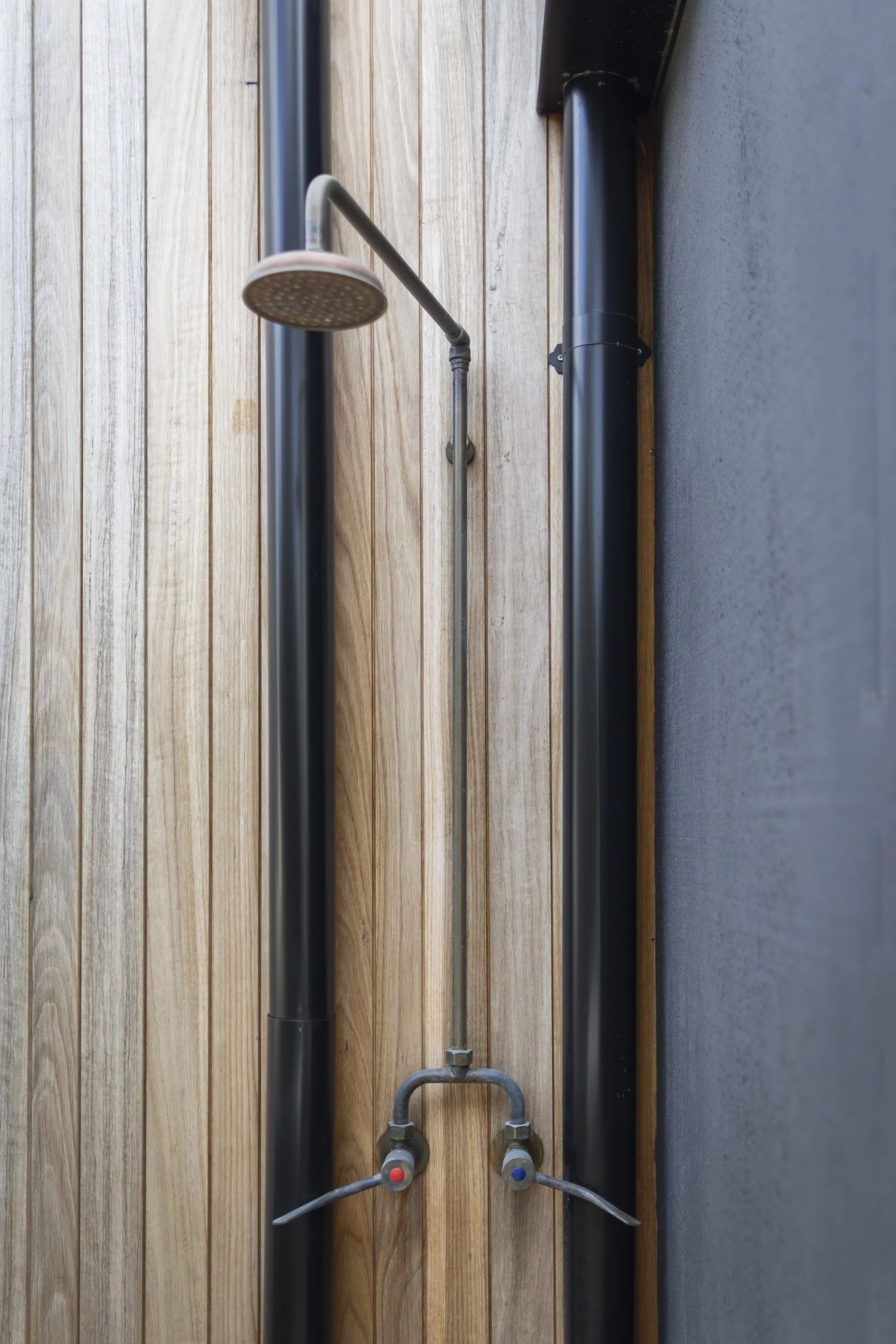 Metal pipe with a shower head attached, mounted on a wooden wall with a black pipe and electrical wiring nearby.