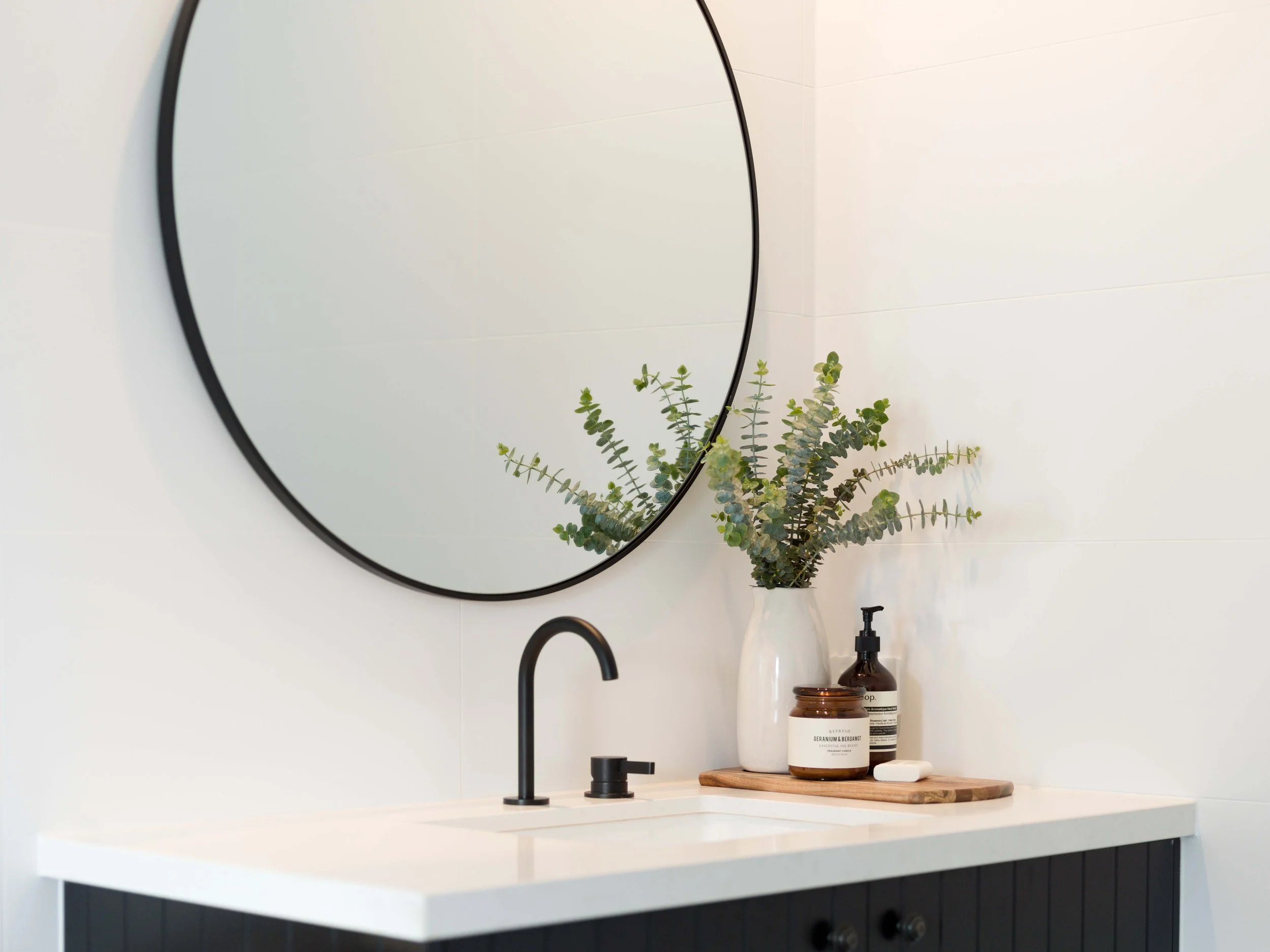 A bathroom vanity with a black curved faucet, a small potted eucalyptus plant, brown and white skincare jars, soap, and a round mirror on a white countertop and wall.