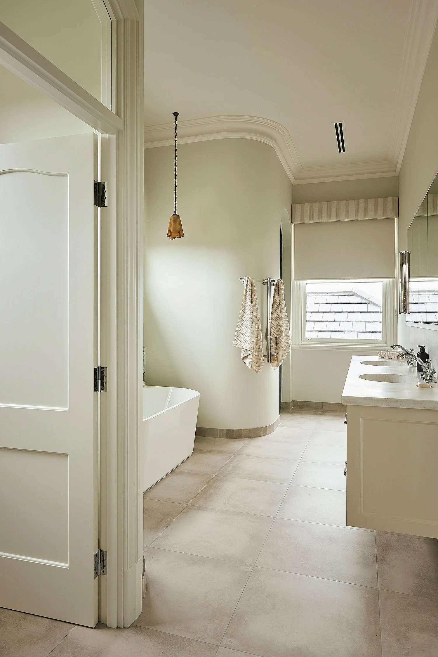 Bright bathroom with a freestanding bathtub, a double sink vanity, a window with blinds, a towel rack with towels, and a decorative ceiling with crown molding.