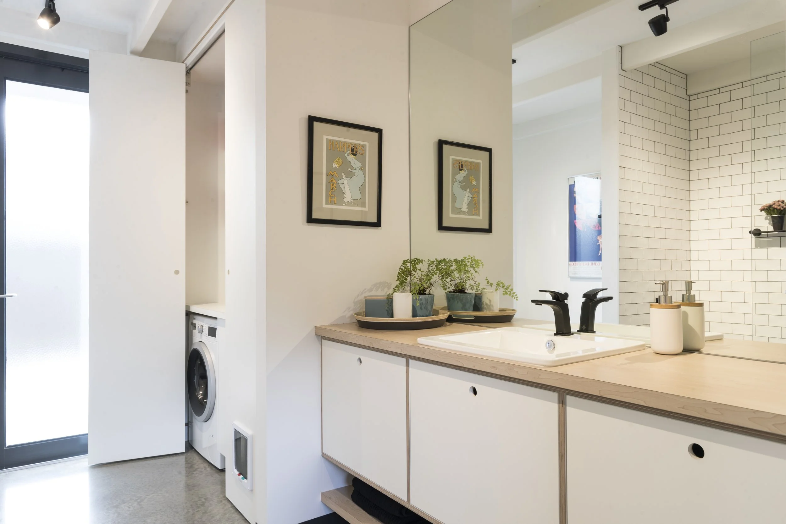 A modern bathroom with a white vanity, black faucet, and a large mirror. On the countertop, there are potted plants and soap dispensers. The wall is made of white subway tiles, and part of a washing machine is visible under the countertop. Artwork ha