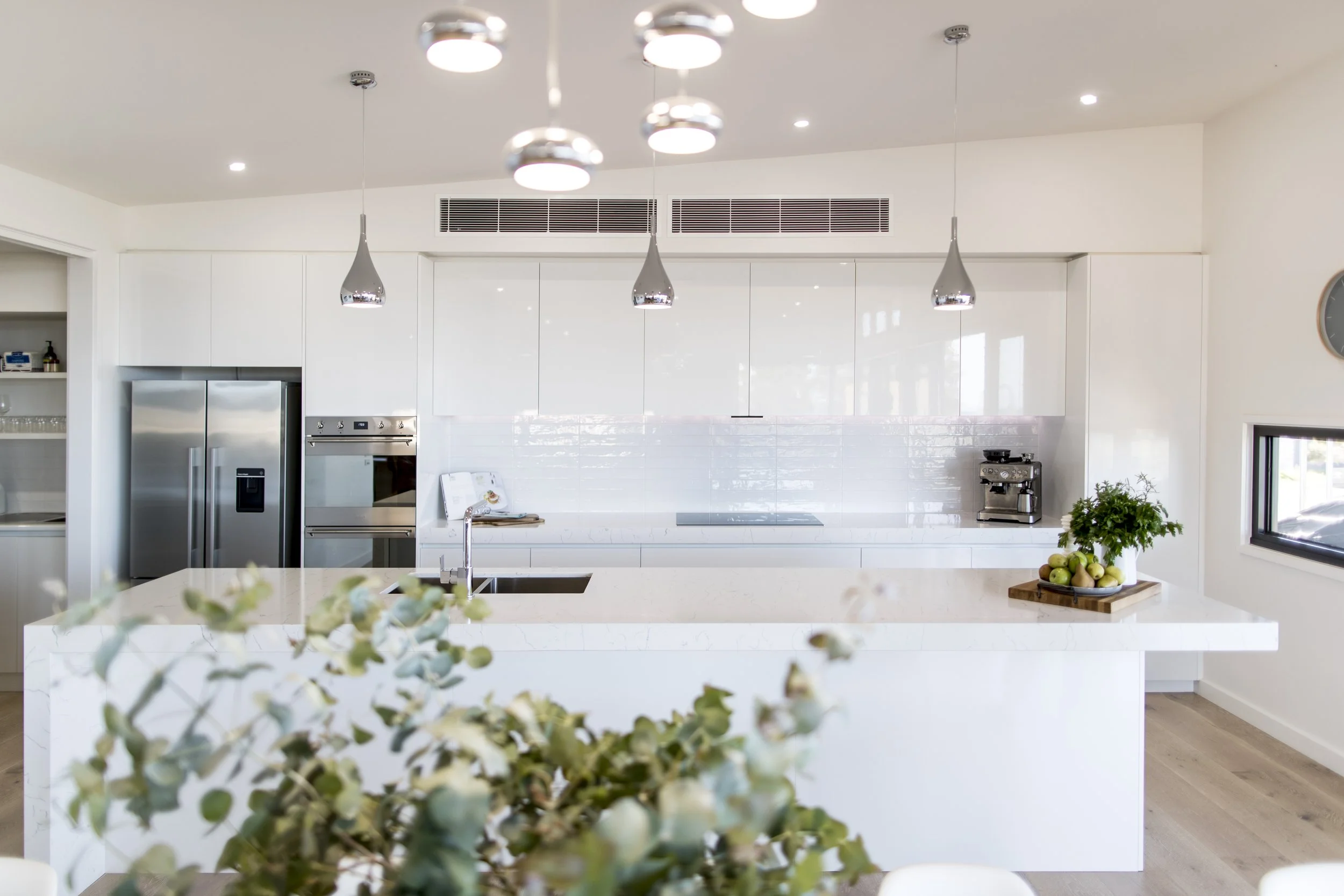 Modern white kitchen with hanging pendant lights, stainless steel refrigerator, and a kitchen island with a bowl of green apples and a potted plant.