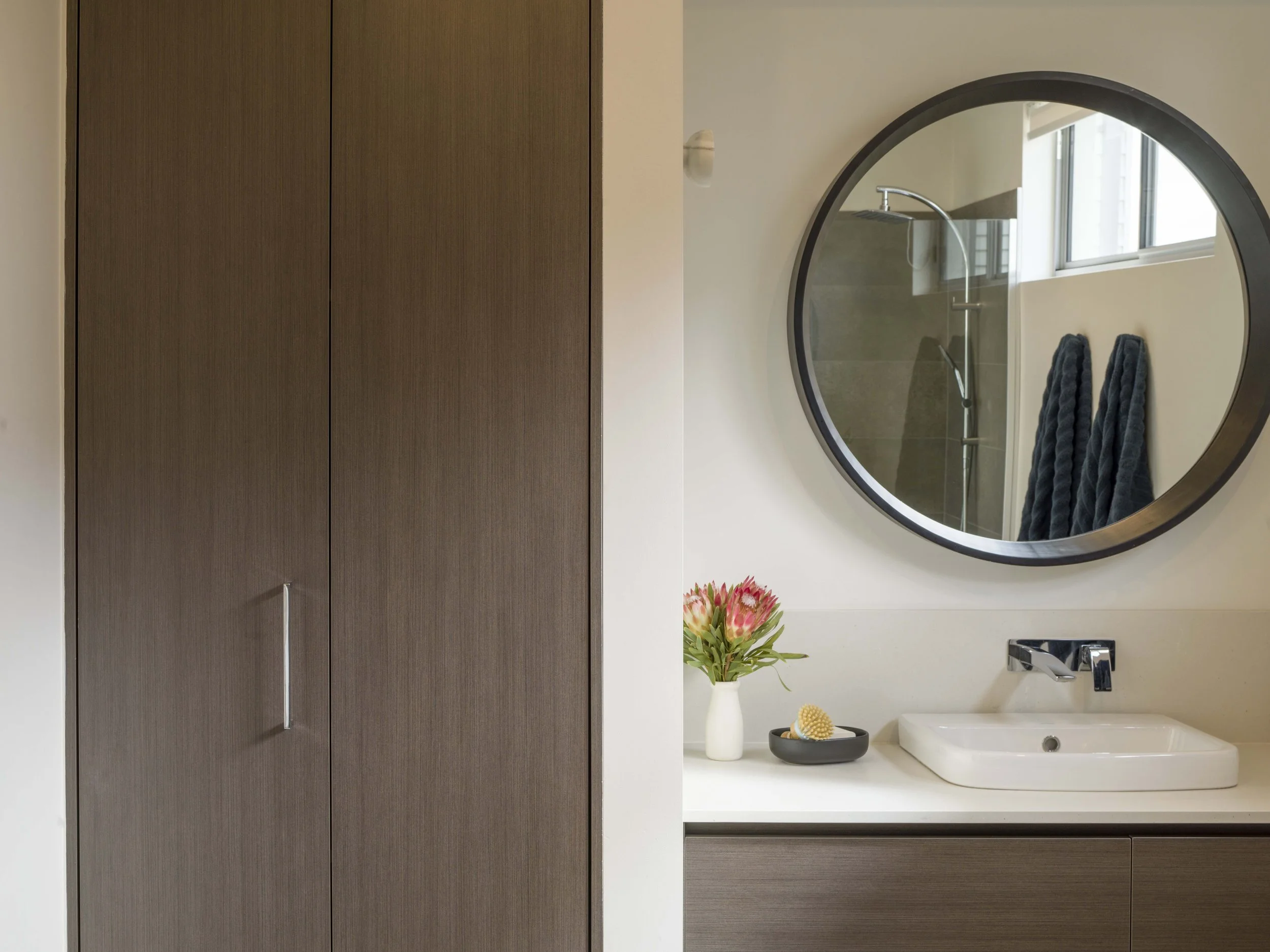 Bathroom with a large round mirror, a bathroom sink, a faucet, a potted plant, and a black tray with a bath brush on the countertop. The bathroom has window lighting and modern decor.