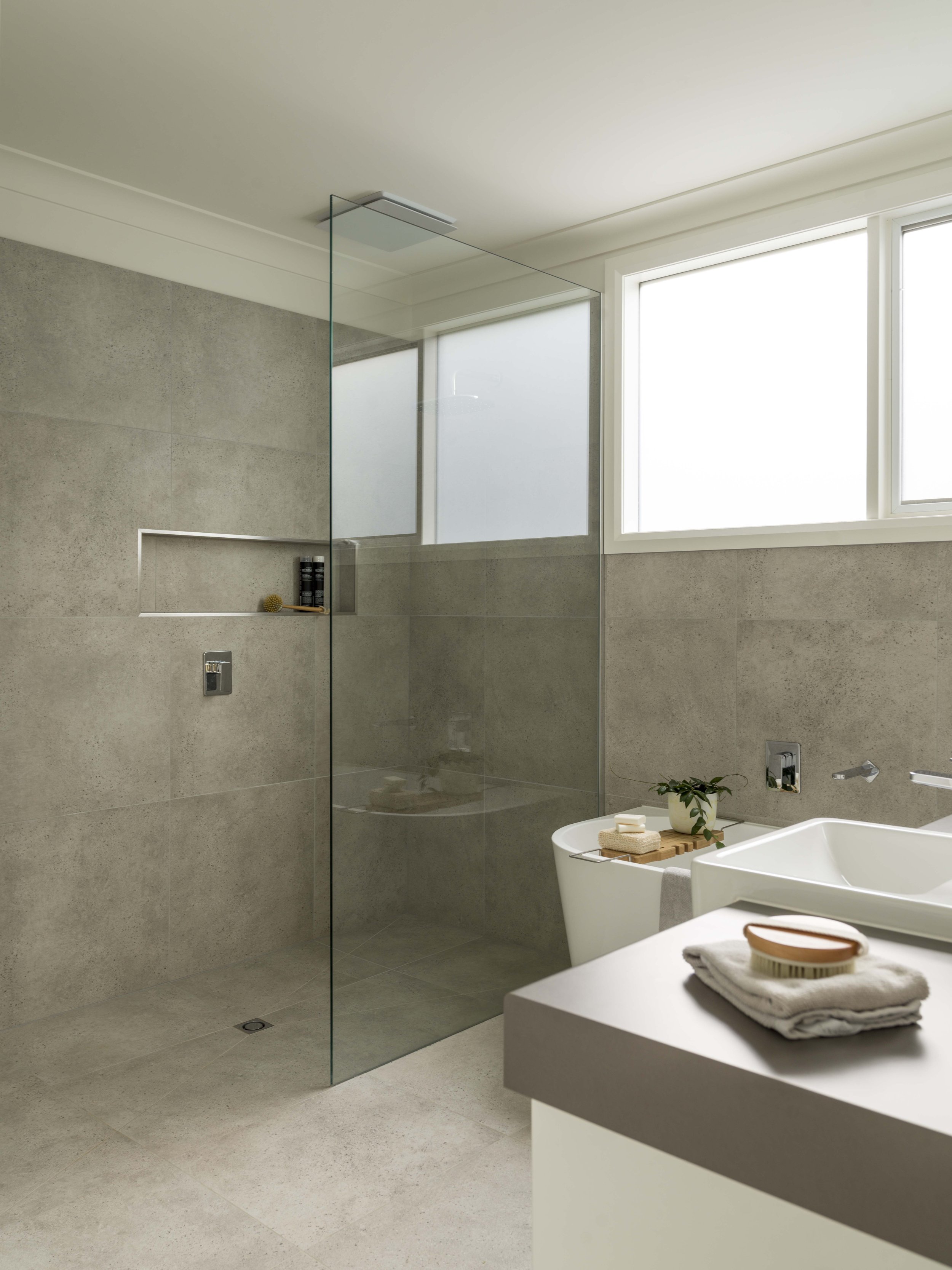 Modern bathroom with a large window, beige tiled walls, a glass shower partition, a white bathtub, and a white countertop with folded towels and a wooden tray, decorated with a small plant.