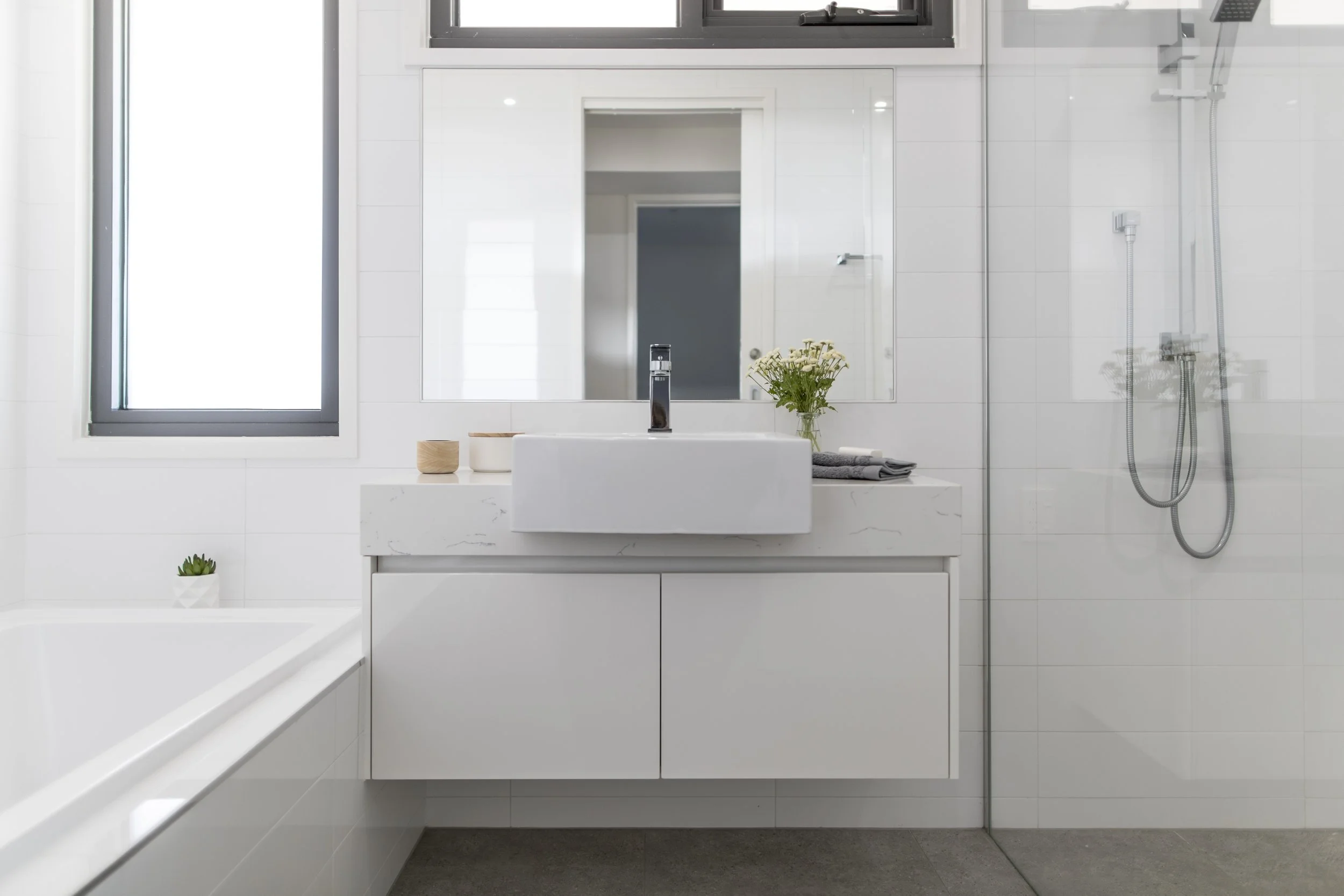 A modern, minimalist bathroom with white tiled walls, a large window, a white vanity with a sink, a mirror, a small potted plant, towels, and a walk-in shower with a glass wall and a handheld showerhead.