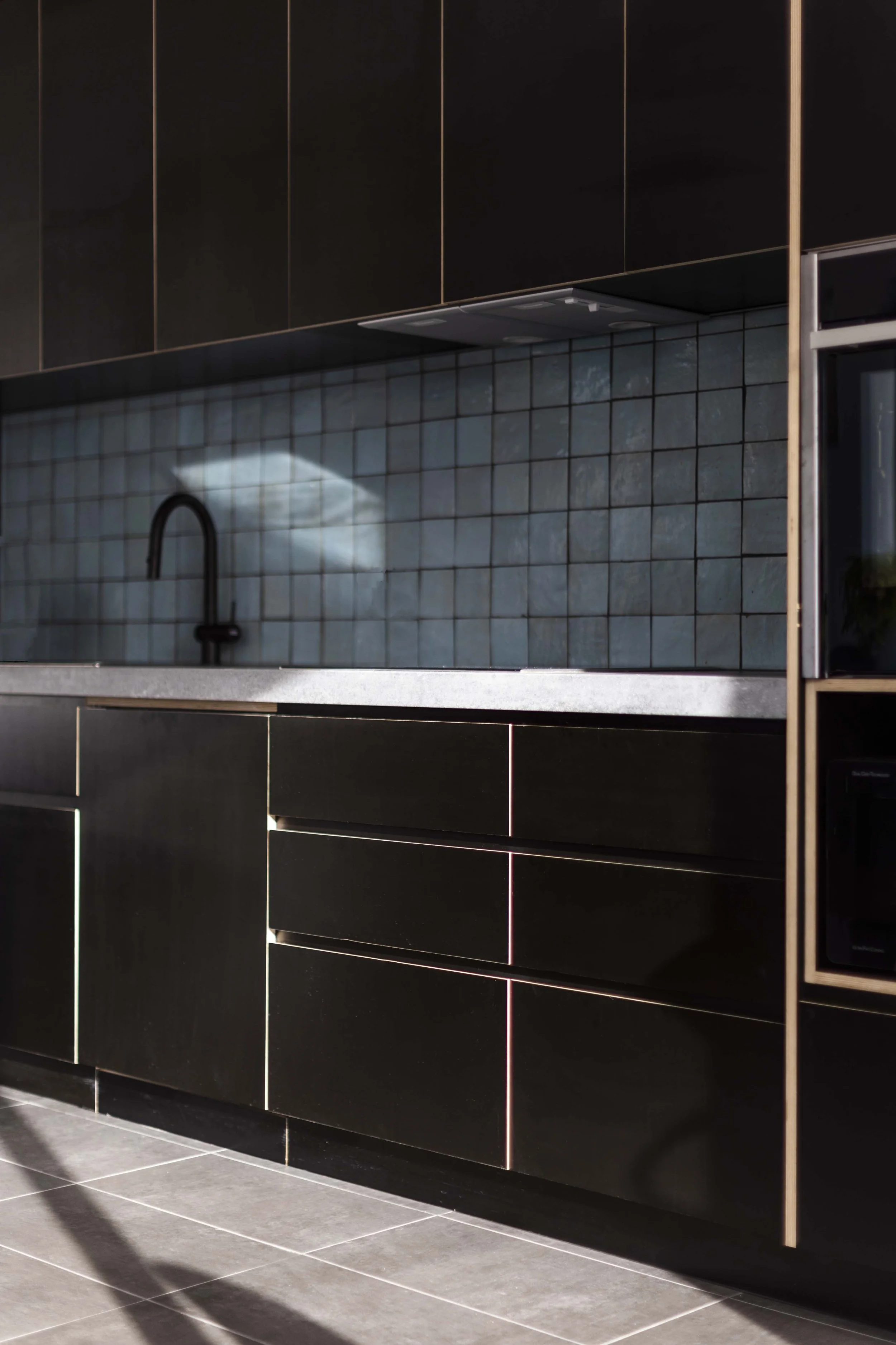 Modern dark cabinetry in a kitchen with a tiled backsplash, black sink faucet, and shadowed floor tiles.