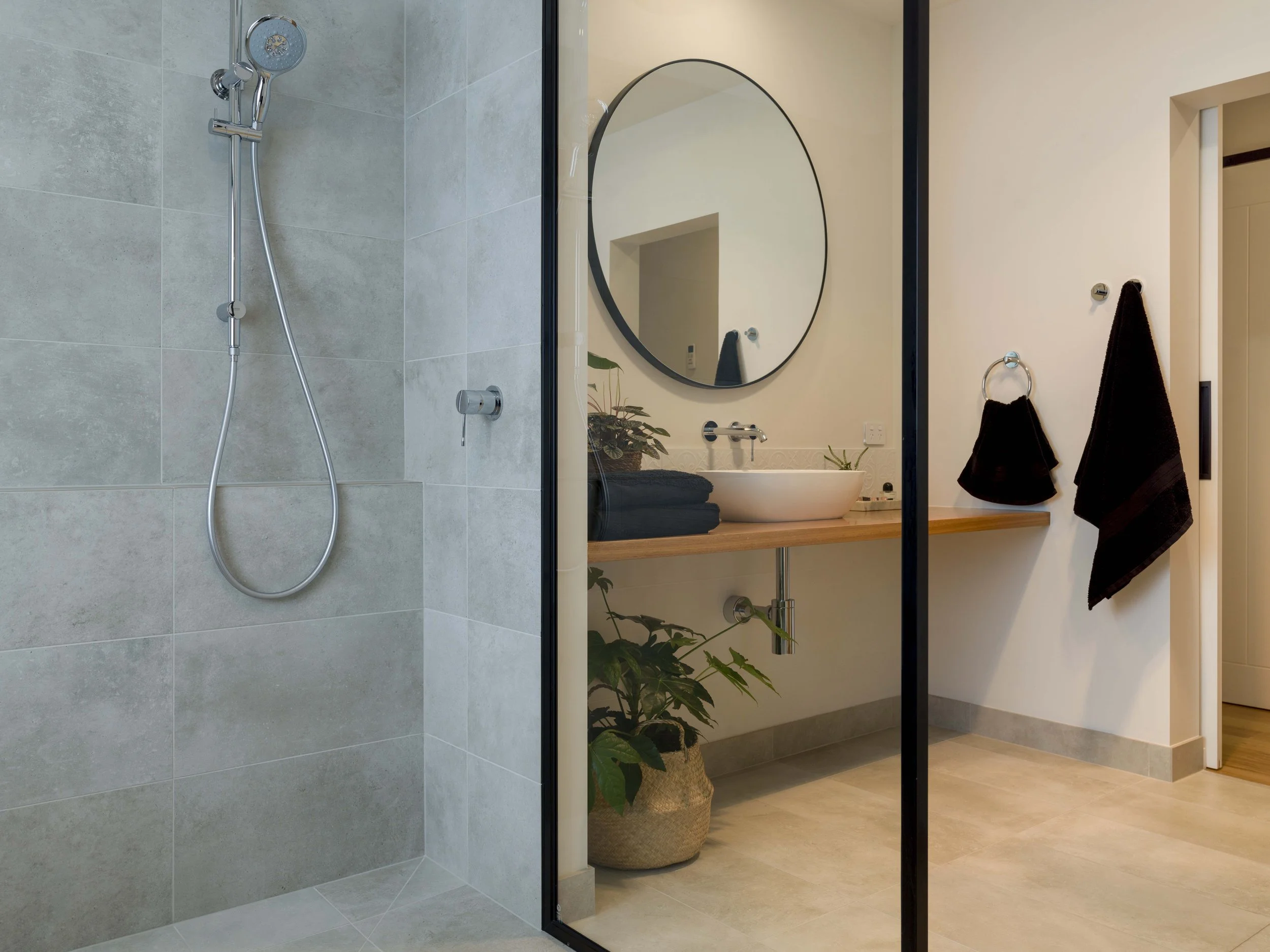 Modern bathroom with a walk-in shower with gray tiles, a large round mirror, a sink with black towels, and a potted plant, with a glimpse of a room with wooden flooring.