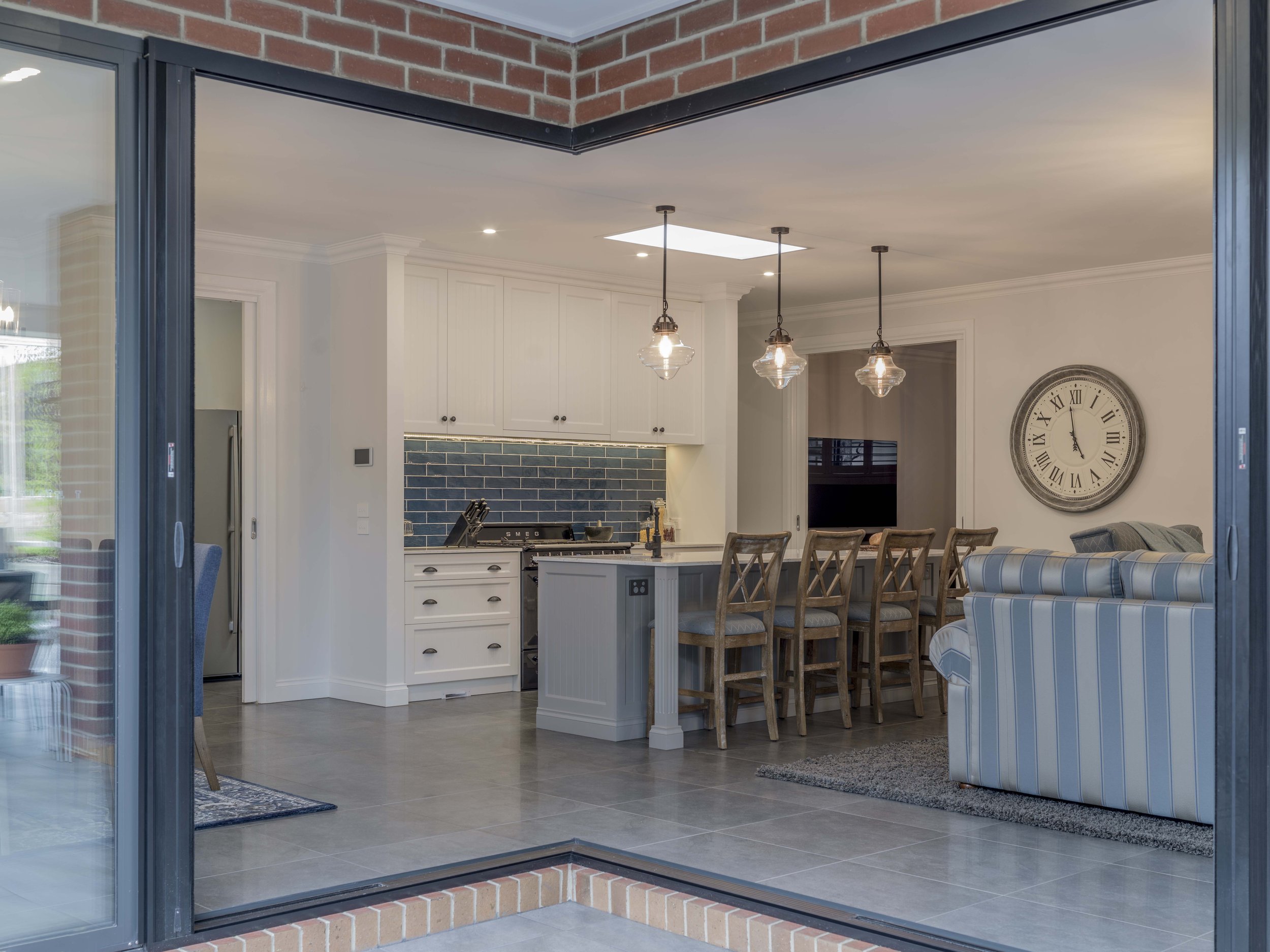 View through sliding glass doors into a kitchen and dining area with white cabinetry, a blue tiled backsplash, hanging pendant lights, a large wall clock, and a striped sofa.