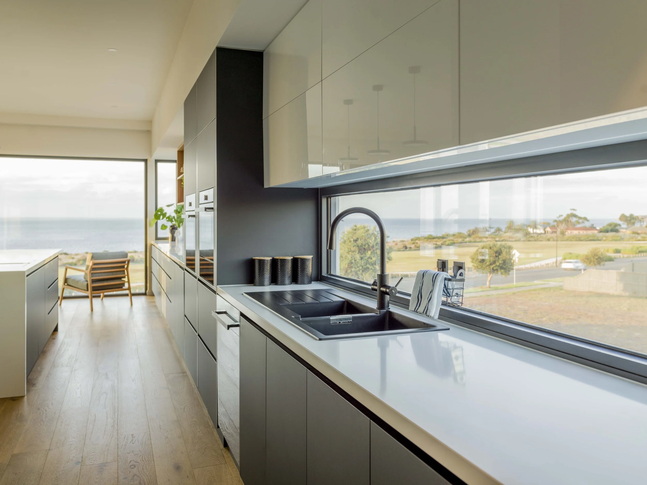 Modern kitchen with white cabinets and a black countertop, large window with a view of trees and ocean, wooden flooring, and a dining area with wooden chairs.