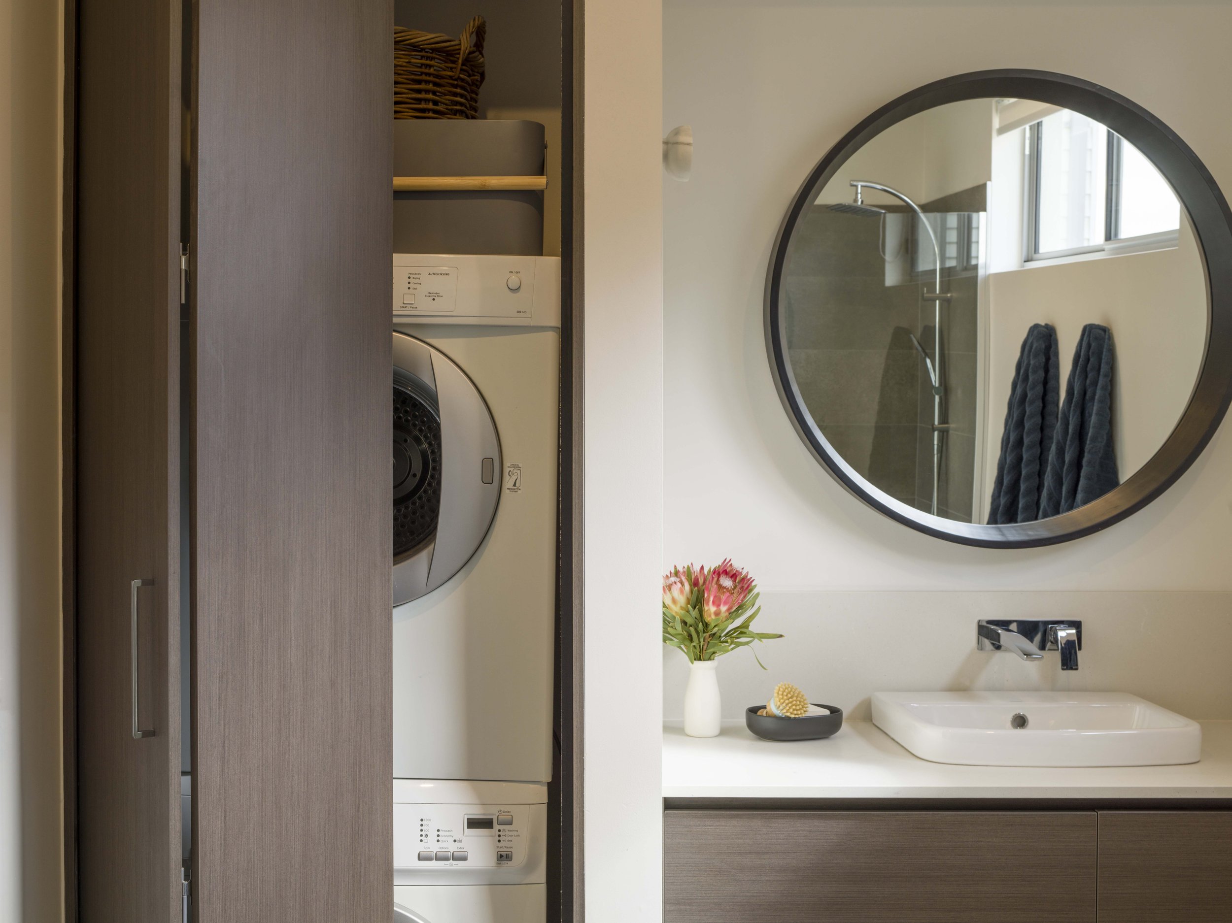 A bathroom with a laundry area, featuring a washing machine behind a wooden sliding door, a round mirror above a sink, a vase with pink flowers, towels, and bathroom toiletries.