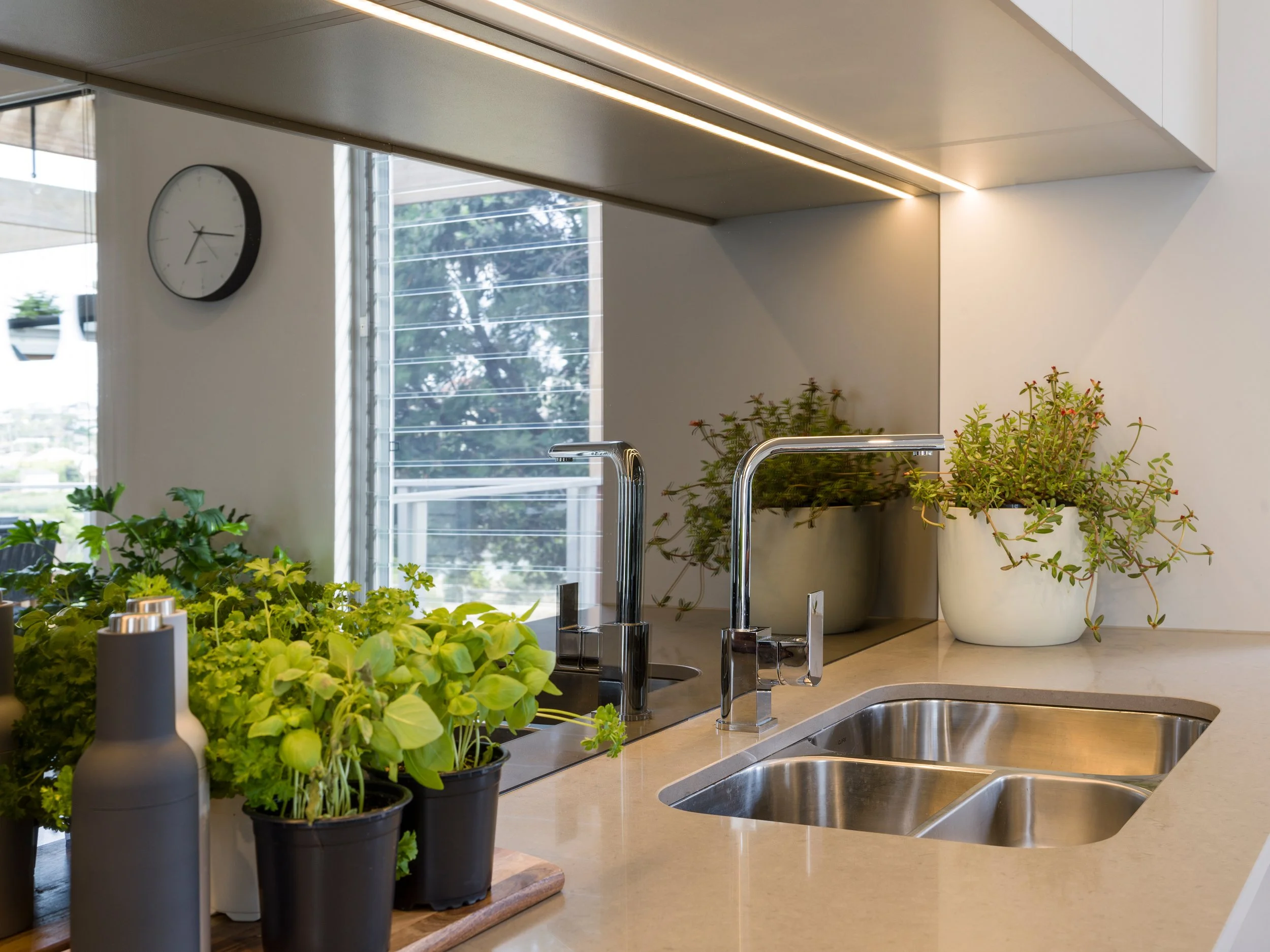 A modern kitchen counter with various potted herbs and plants near a sink, a window with blinds, a wall clock, and overhead lighting.