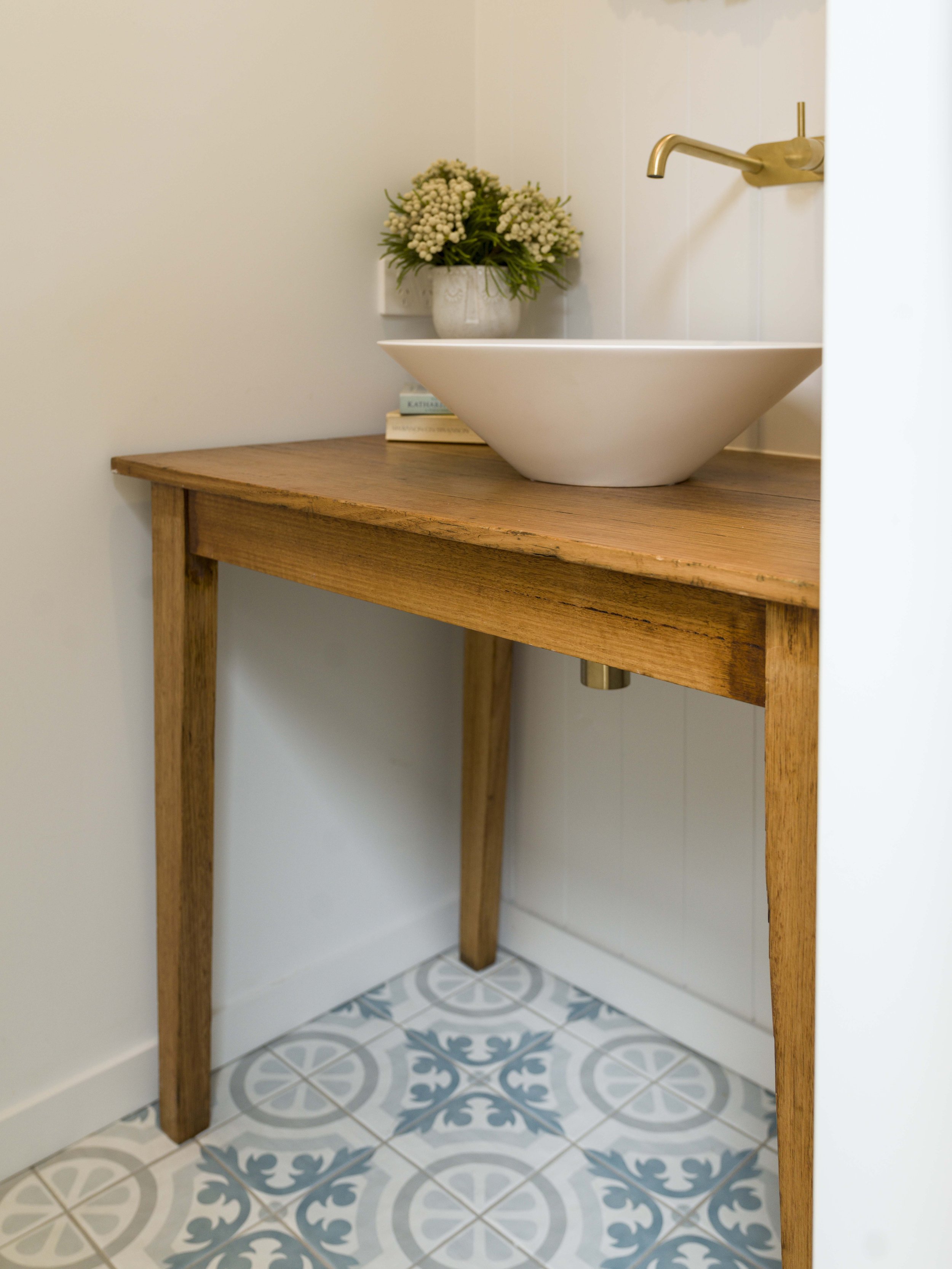 A bathroom sink with a wooden vanity, a faucet, a vase with white flowers, and books on the vanity. The floor has patterned tiles in shades of blue and gray.
