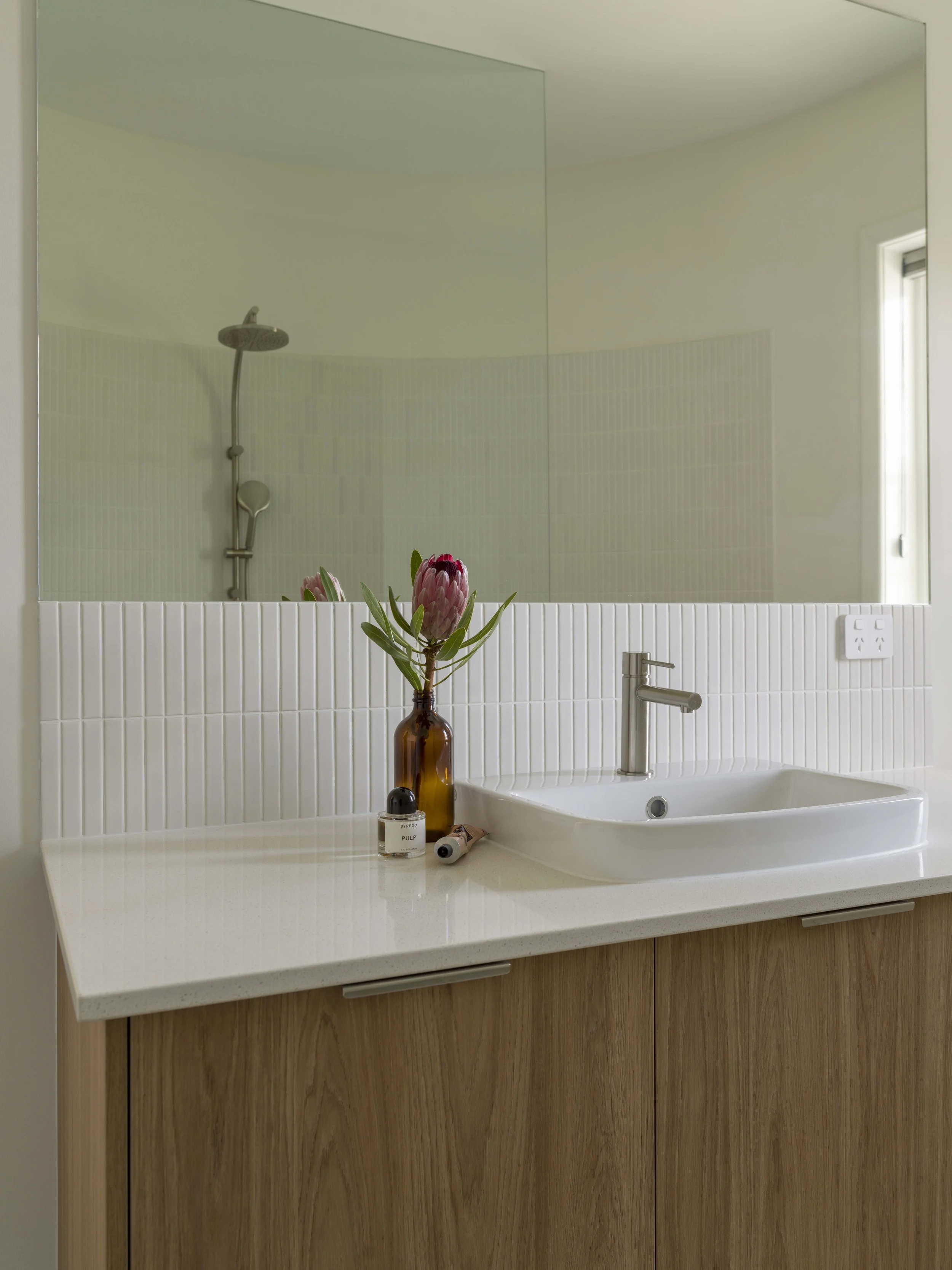 A bathroom vanity with a white sink, a modern faucet, a mirror, a small brown vase with pink protea flowers, and some skincare and beauty products on the counter.