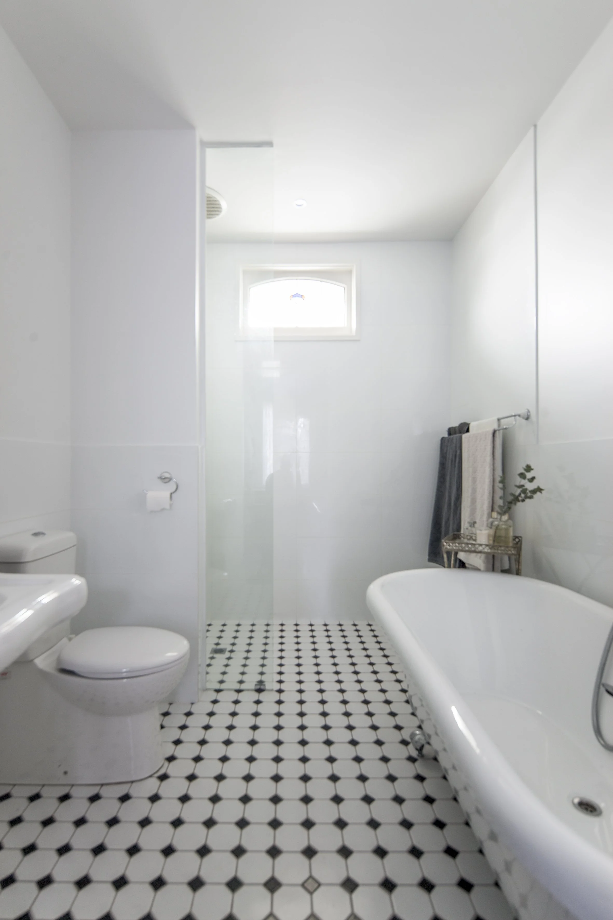 A bathroom with white walls, a clawfoot bathtub, black and white hexagon floor tiles, a white toilet, a small window, and a towel rack with dark towels.