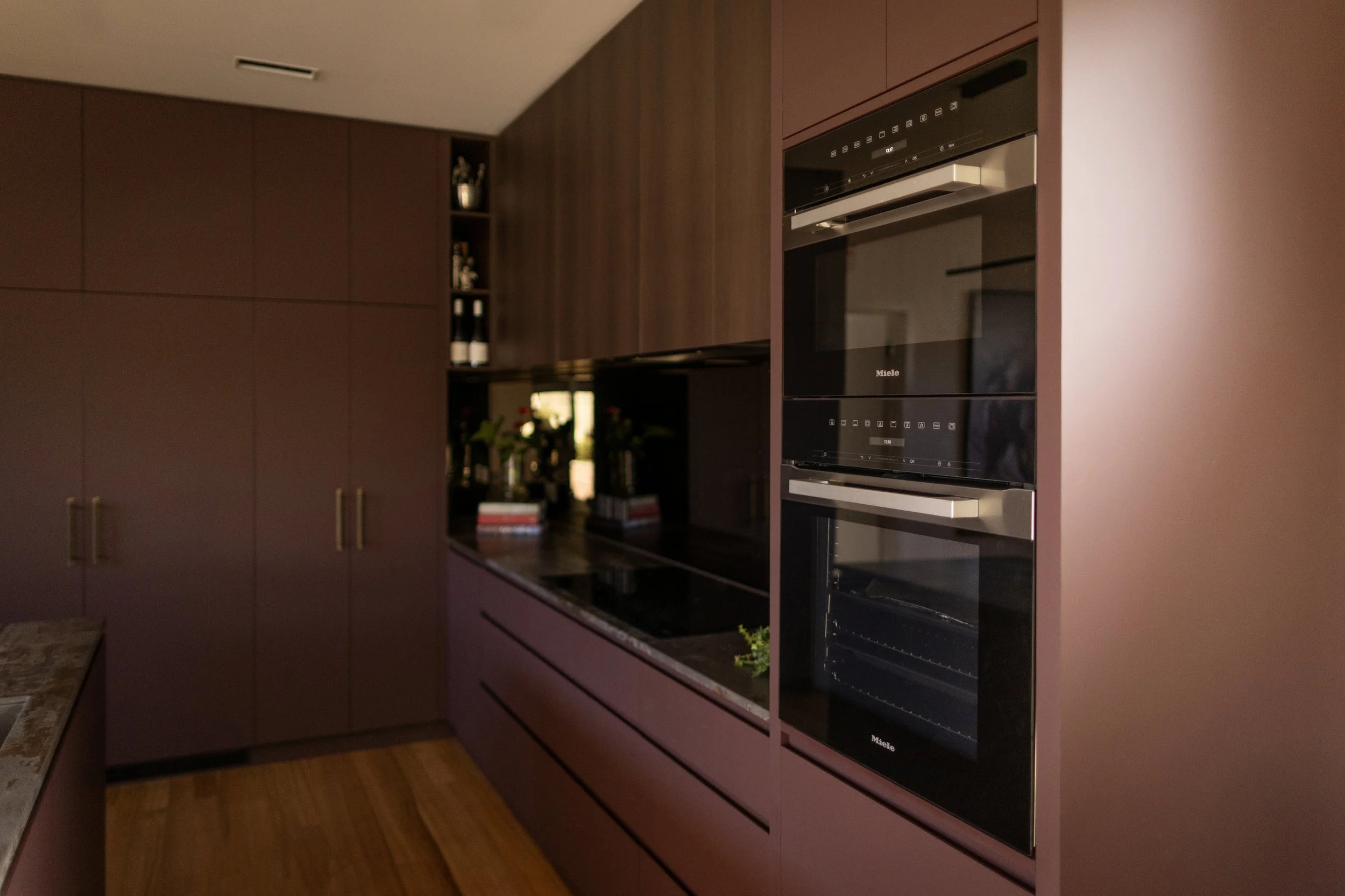 A modern kitchen with purple cabinets and built-in black appliances, including an oven and microwave, with a wooden floor and minimal decor.