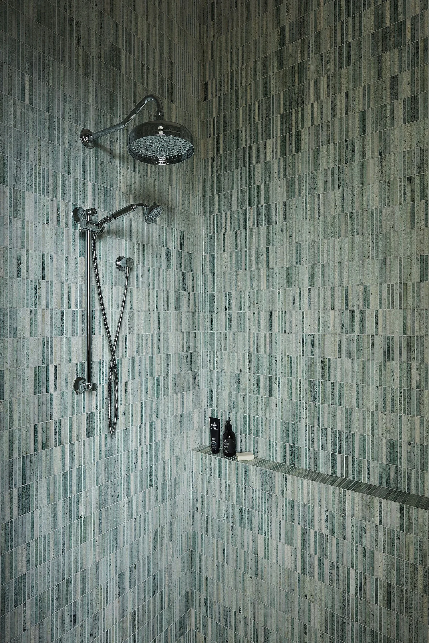 A walk-in shower with green mosaic tile walls, a black rainfall showerhead, a handheld showerhead, and two bottles of toiletries on a built-in shelf.