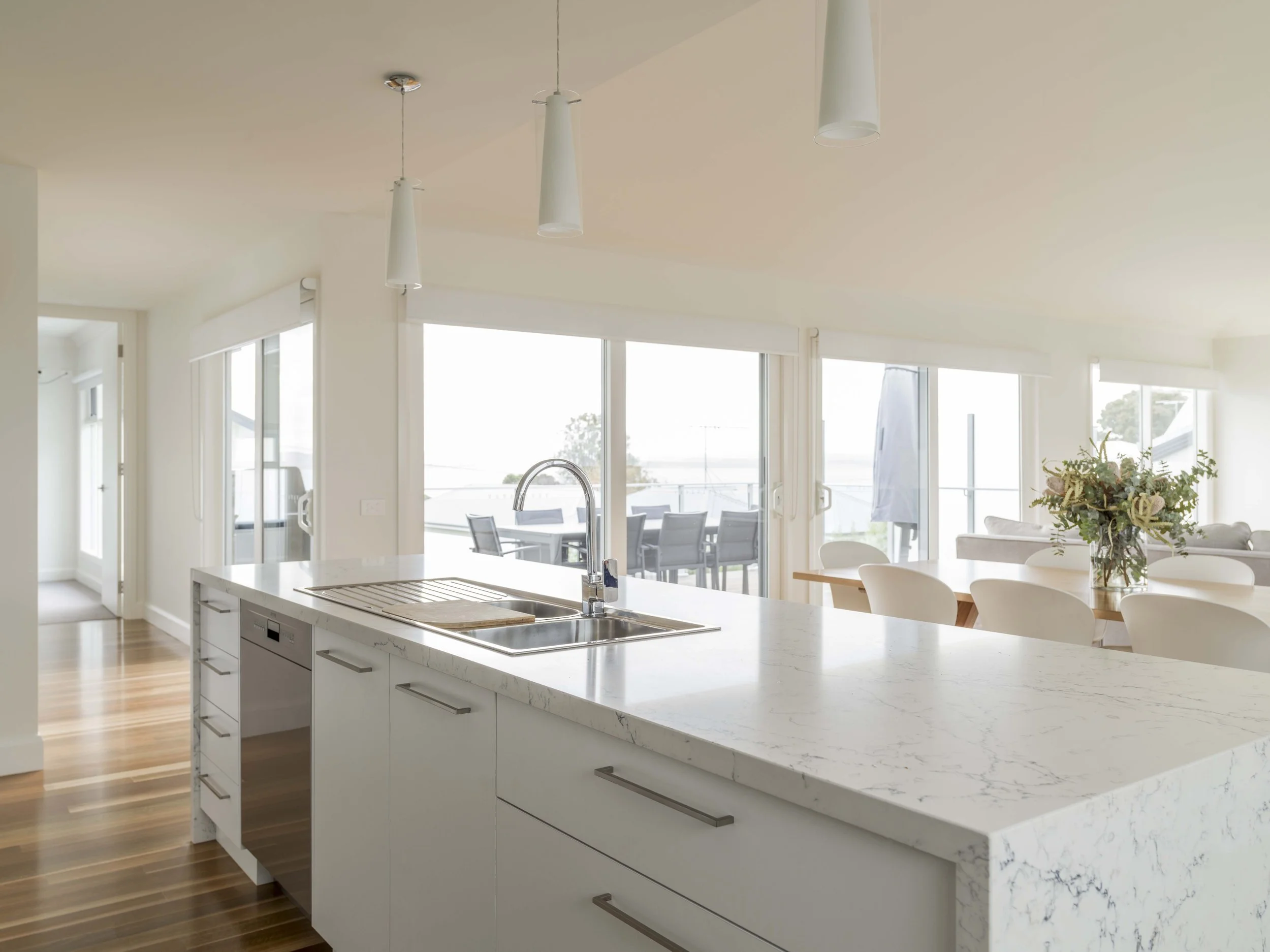 Modern kitchen with a white marble island, pendant lights, large windows, dining area, and a sliding glass door leading to an outdoor patio.